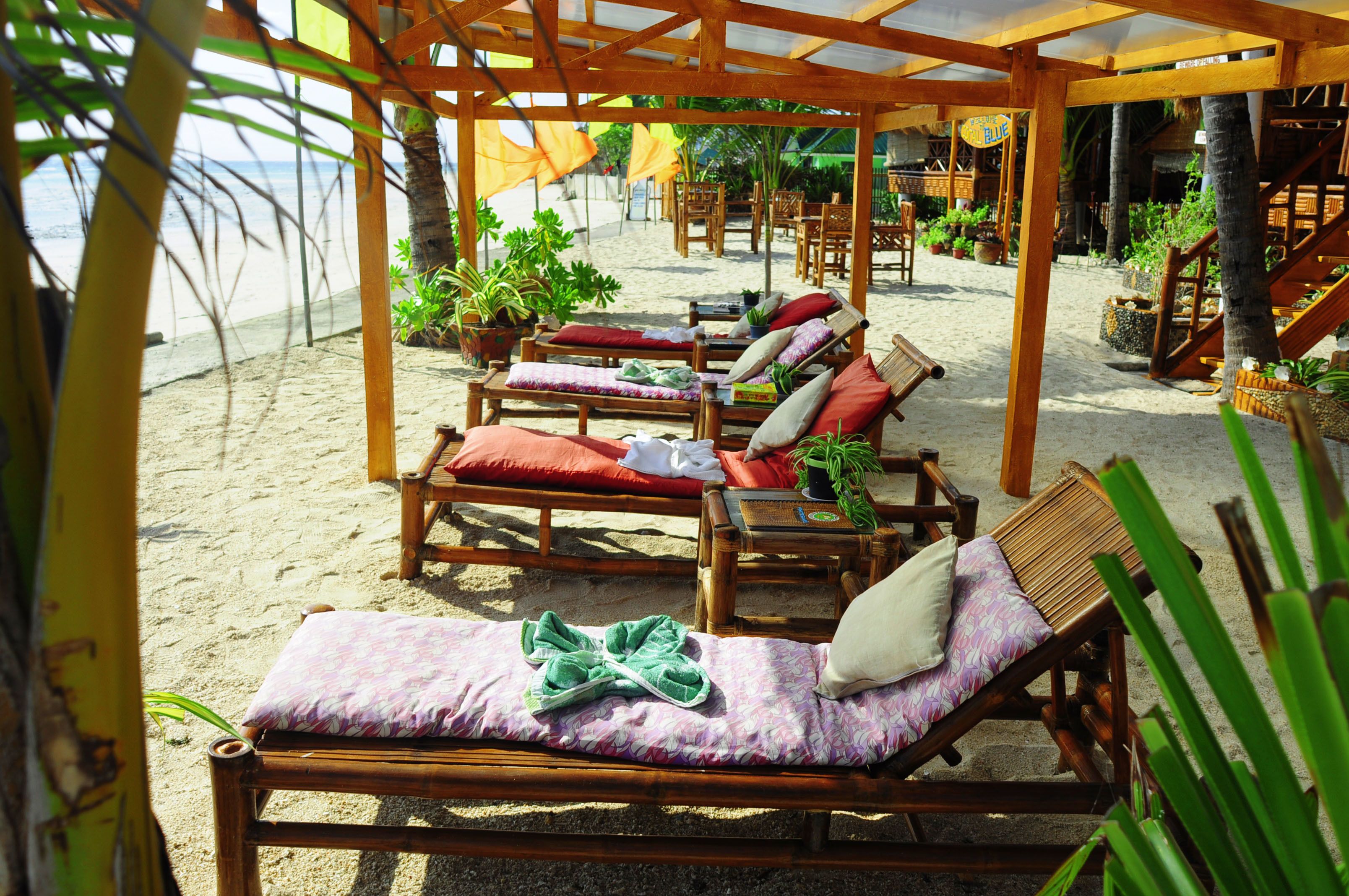 A beachside lounge area with wooden lounge chairs and tables, surrounded by potted plants and umbrellas.