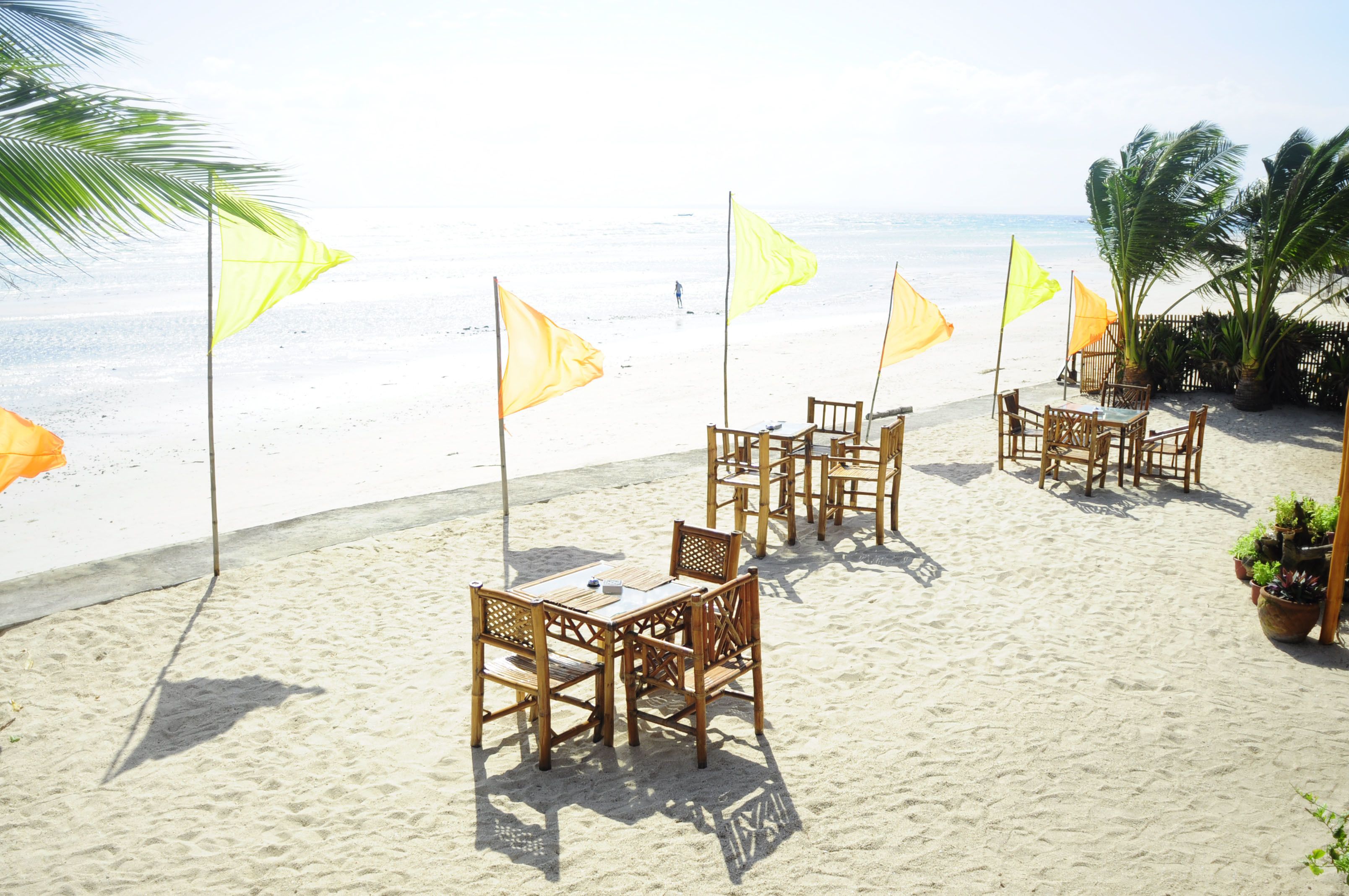 A beachside seating area with wooden tables and chairs, surrounded by yellow flags and palm trees.