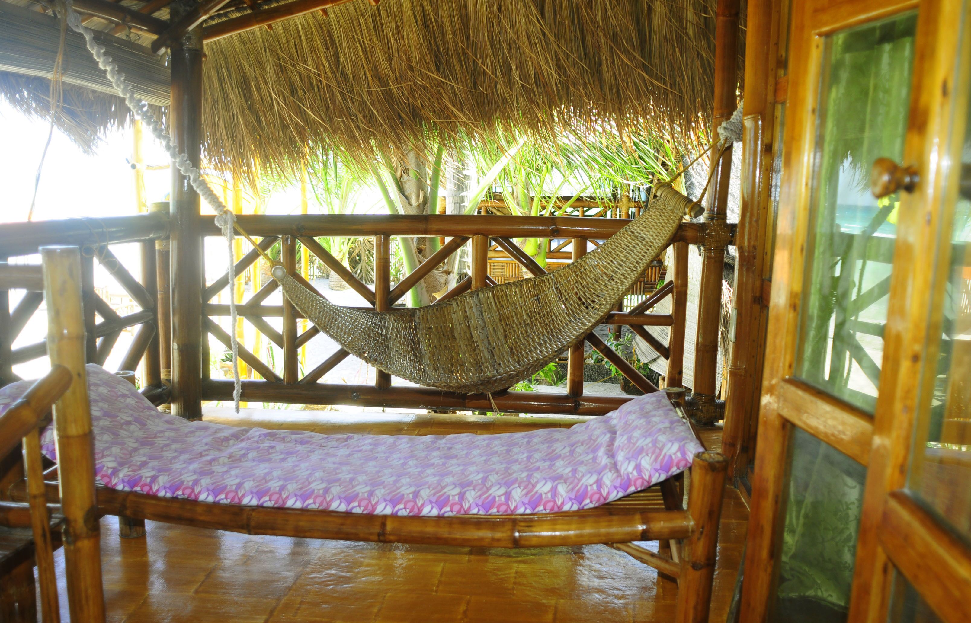 A hammock bed in a wooden structure with a thatched roof, surrounded by bamboo railings and palm trees.