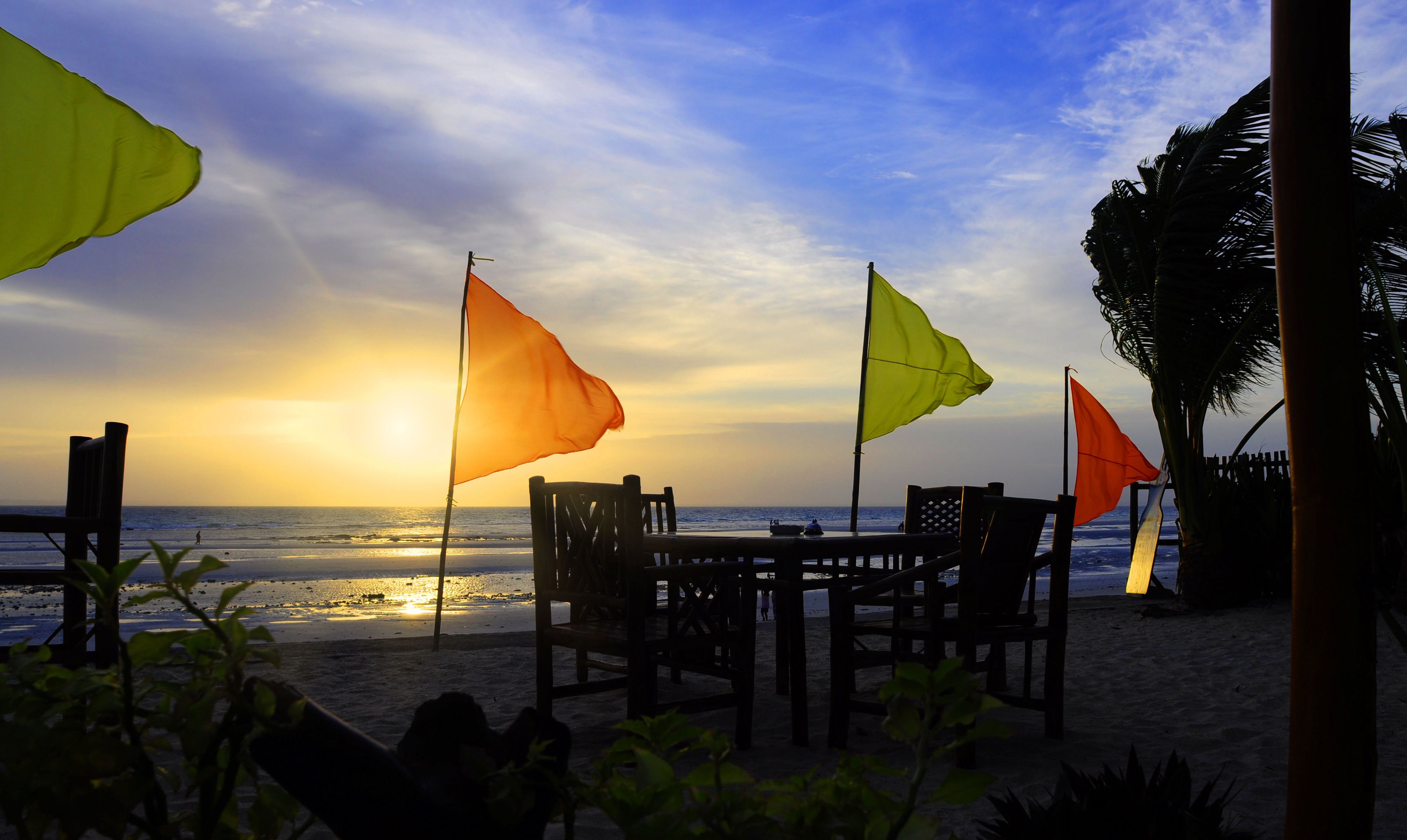 A beachside seating area with tables and chairs, flags, and a view of the ocean at sunset.