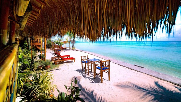 A beachside lounge with a thatched roof, wooden furniture, and a view of the ocean. - The Coral Blue Oriental Beach Villas & Suites (Santa Fe)