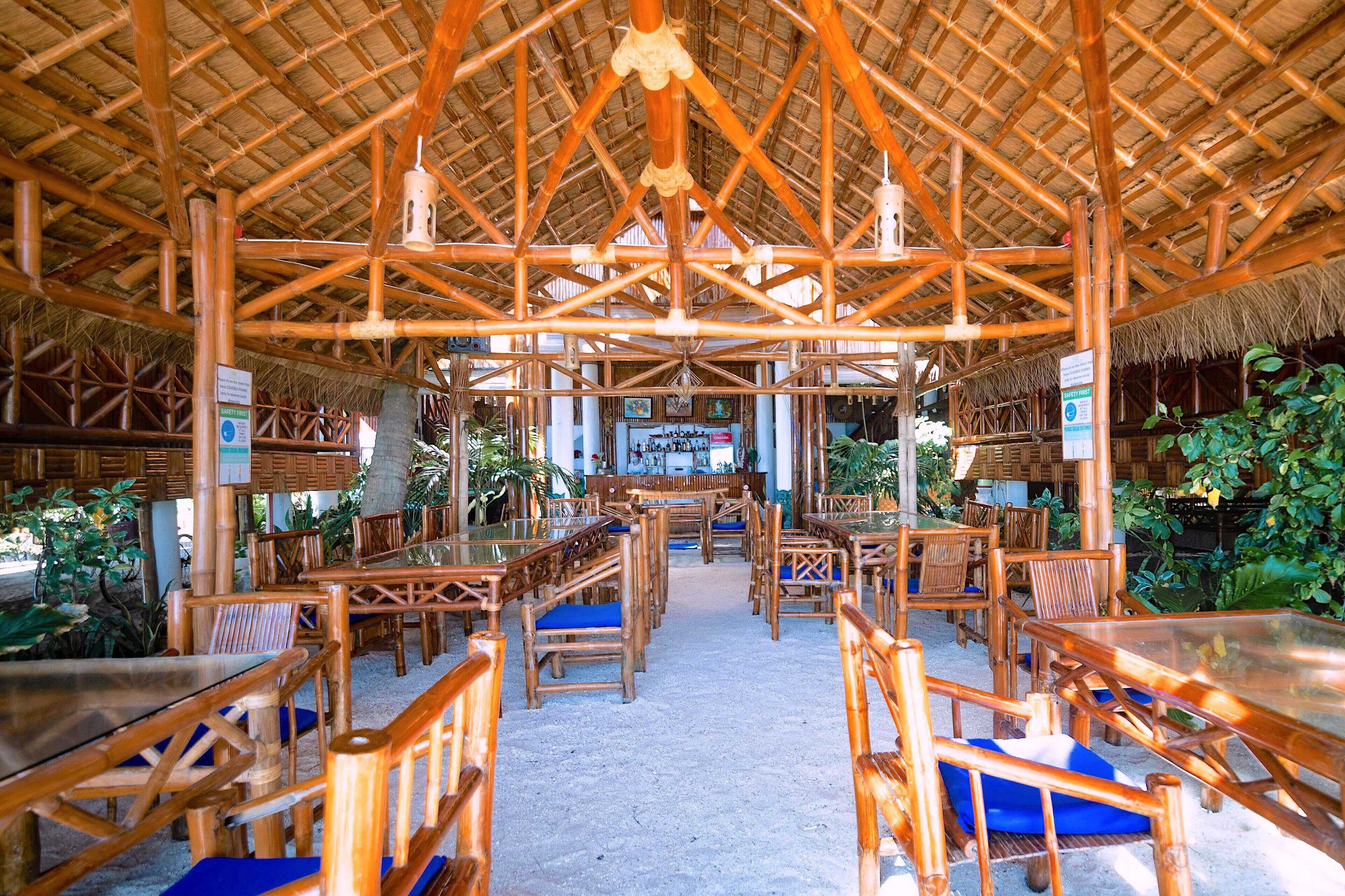 A bamboo-framed indoor dining area with tables and chairs, a thatched roof, and tropical plants.