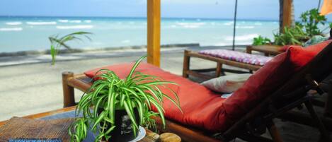 Un salón junto a la playa con vista al océano, una mesa de madera con una planta en maceta y cojines rojos en sillones reclinables.