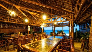 An outdoor dining area with bamboo furniture and a glass table, overlooking the beach. - The Coral Blue Oriental Beach Villas & Suites (Santa Fe)