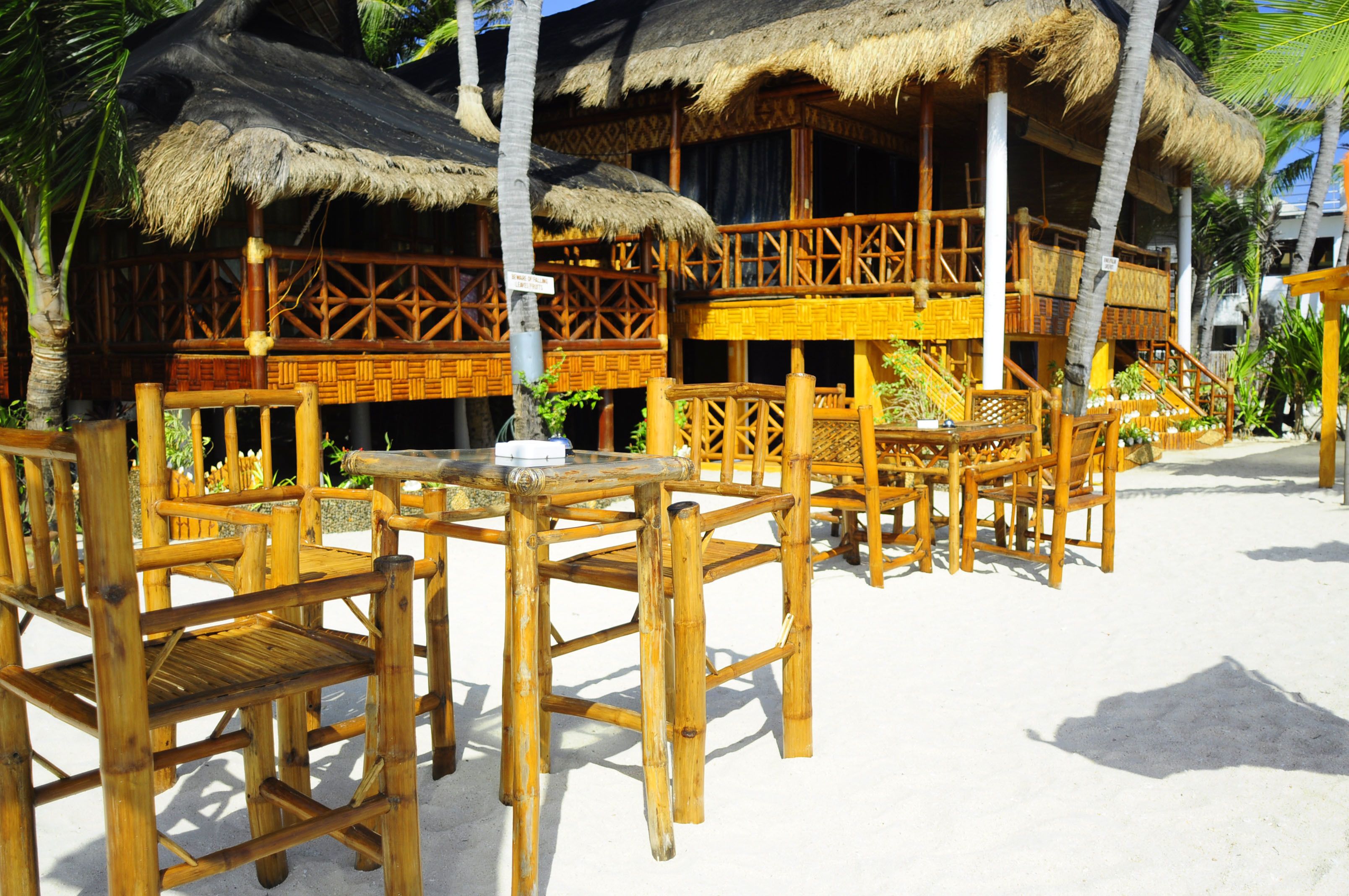 A beachside restaurant with thatched-roof structures, wooden furniture, and a sandy floor.