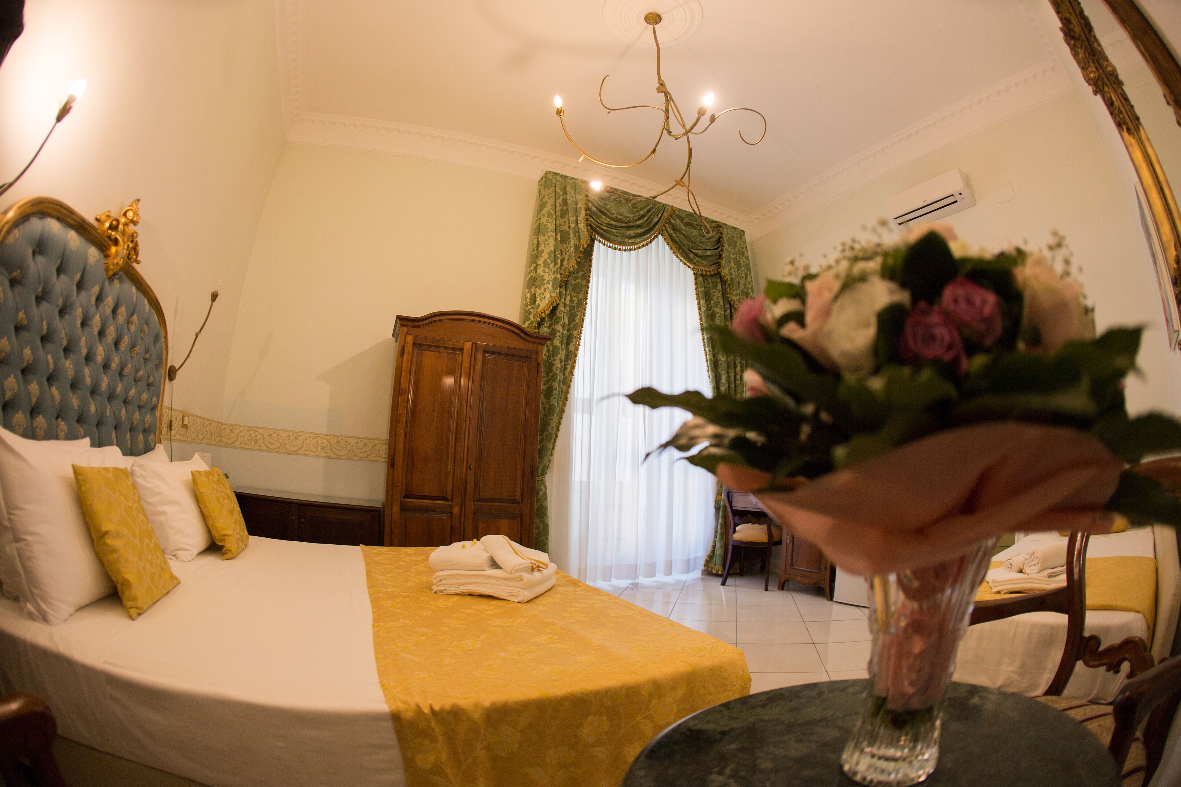 A hotel room with a bed, a wooden wardrobe, a chandelier, and a vase with flowers on a table.