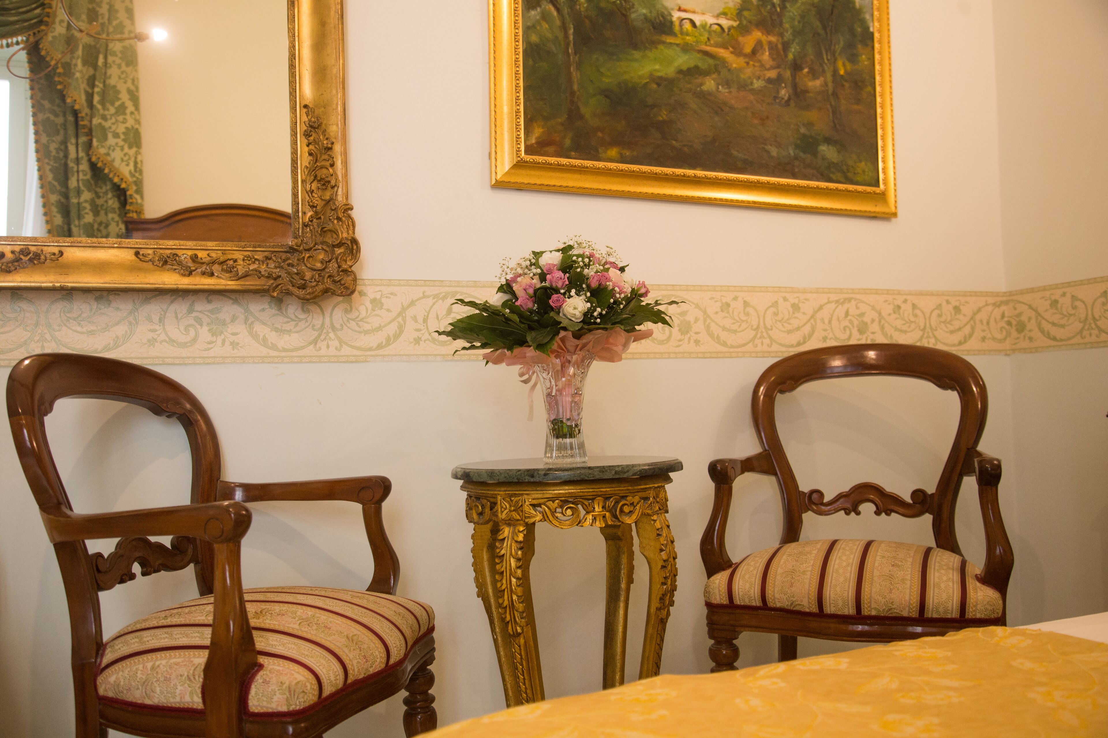 A room with two wooden chairs, a small table with a vase of flowers, and a painting on the wall.