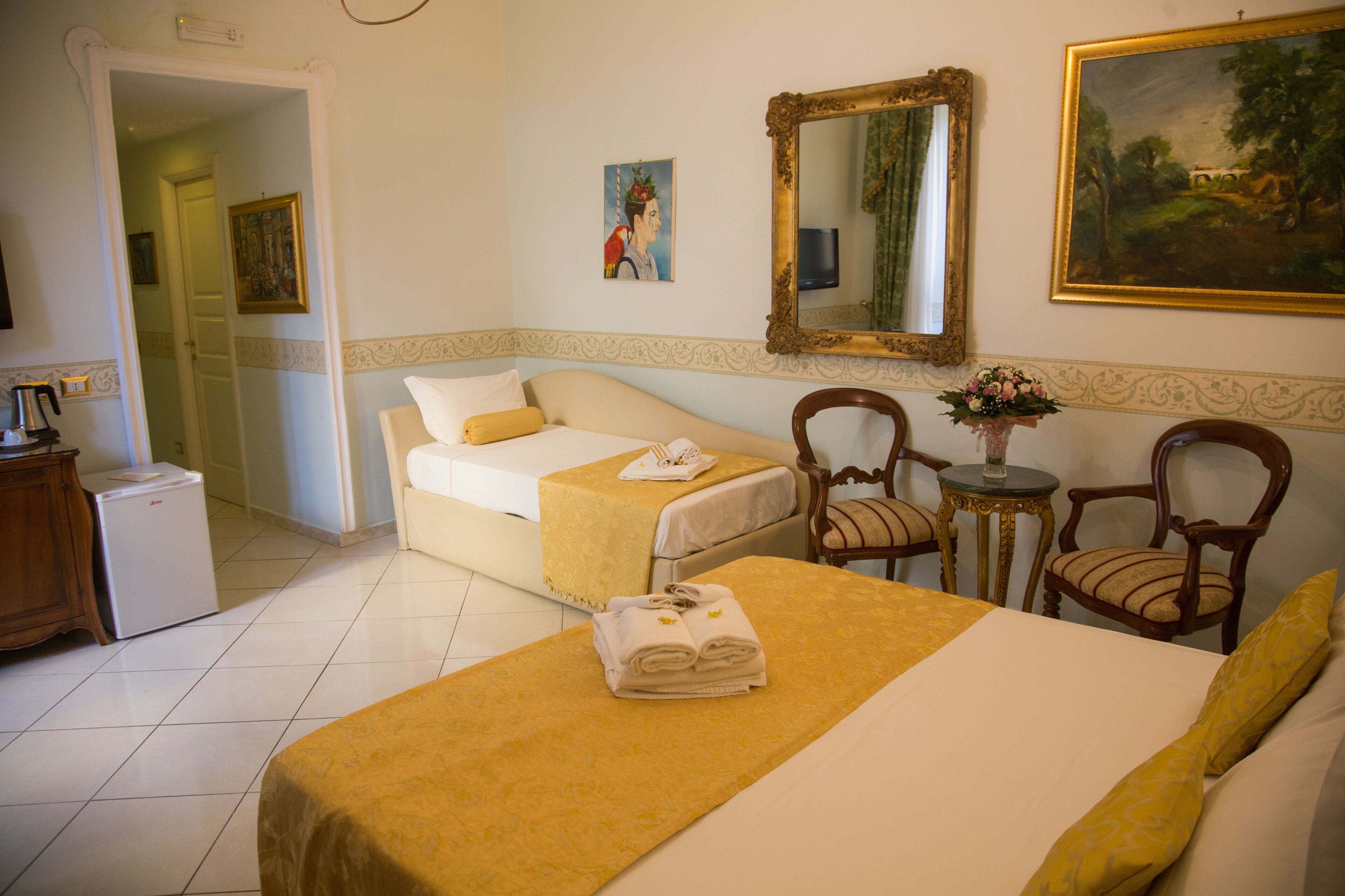 Una habitación de hotel con una cama, una silla, una mesita con un florero y un cuadro en la pared.