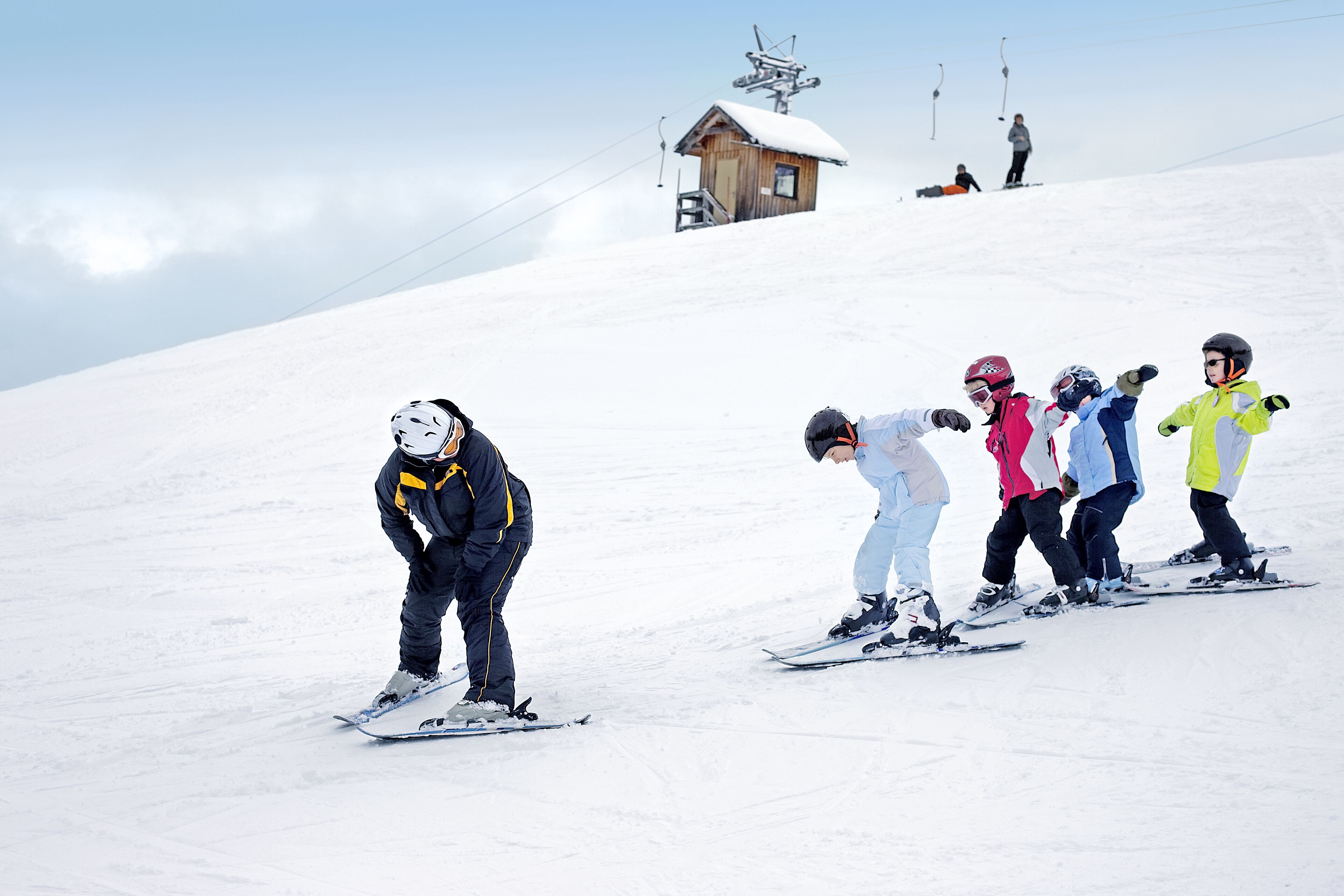 Eine Gruppe von Skifahrern auf einem verschneiten Hang, im Hintergrund eine Holzhütte und eine Seilbahn.