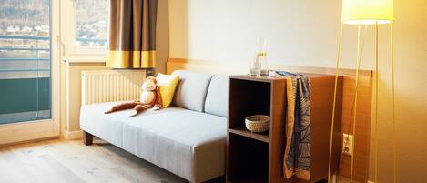 A modern hotel room with a sofa, a small bookshelf, a lamp, and a view of a lake through a window.