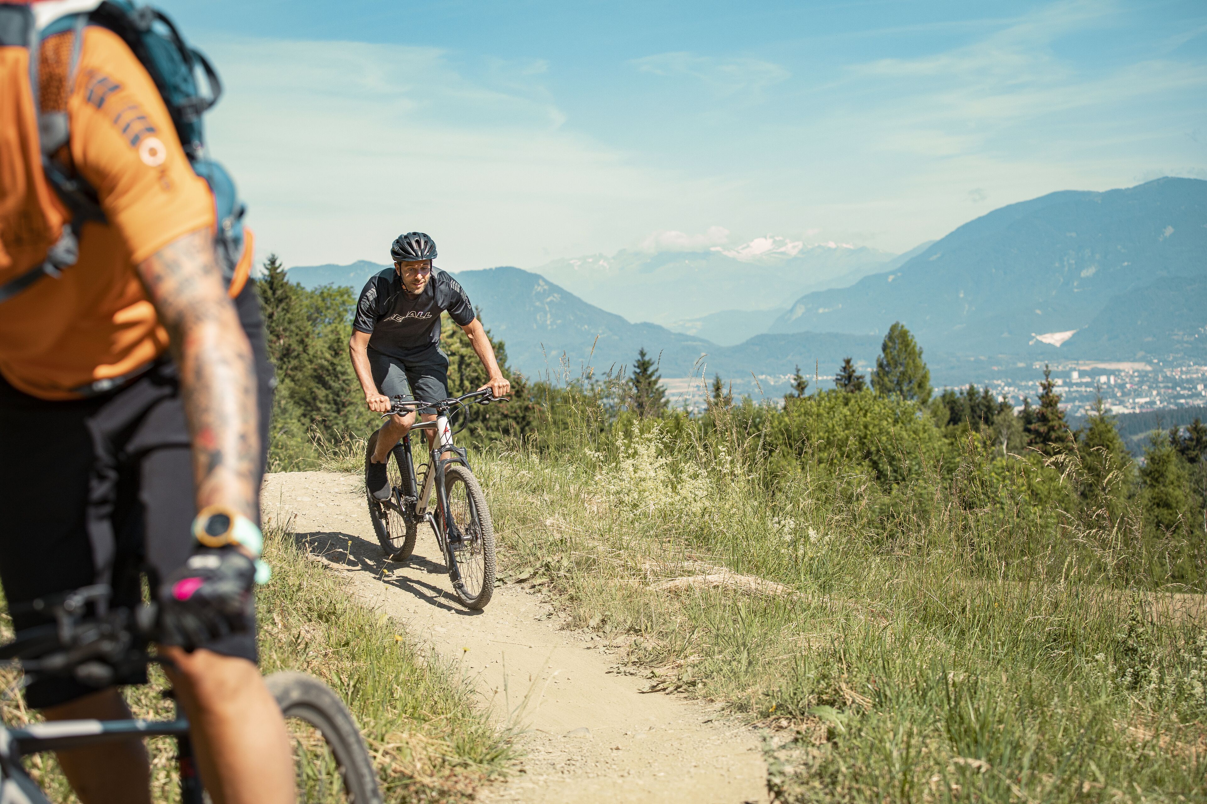A mountain biking trail with a cyclist in the foreground and a scenic mountain backdrop.