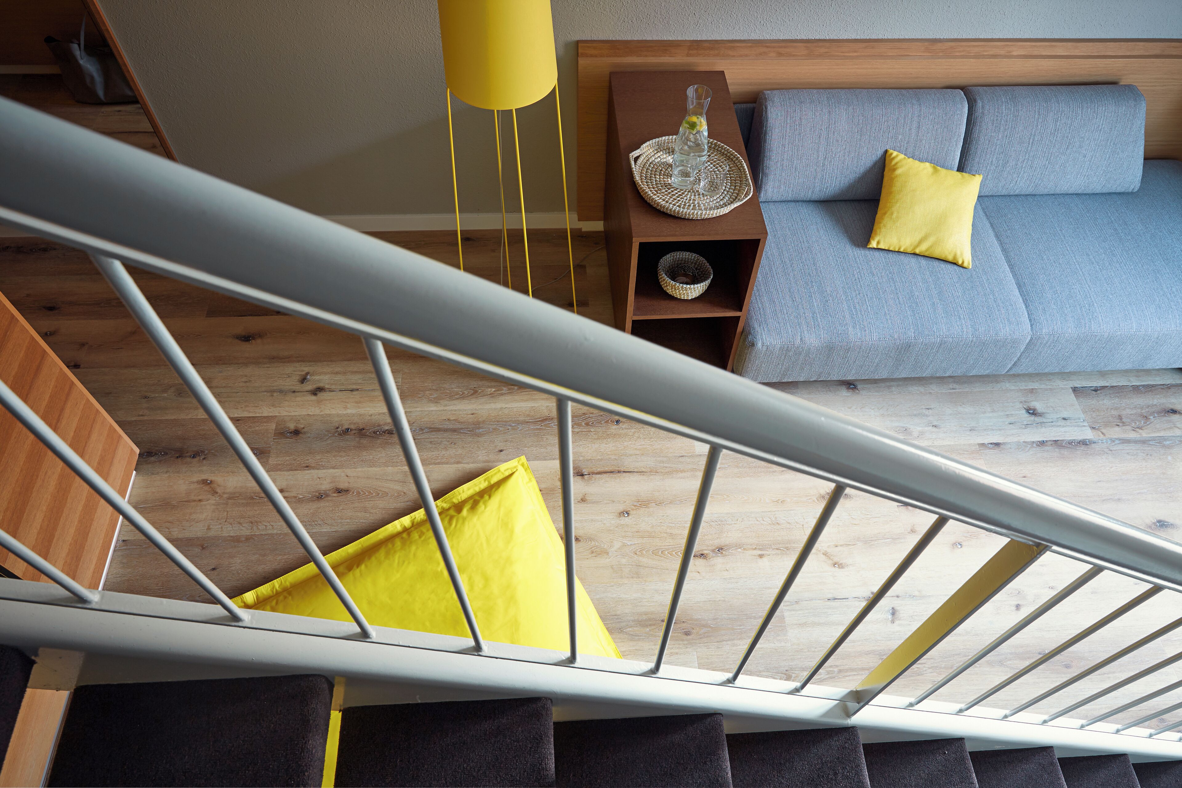 A modern bedroom with a wooden floor, a grey sofa with yellow cushions, a bedside table with a vase, and a yellow lamp.