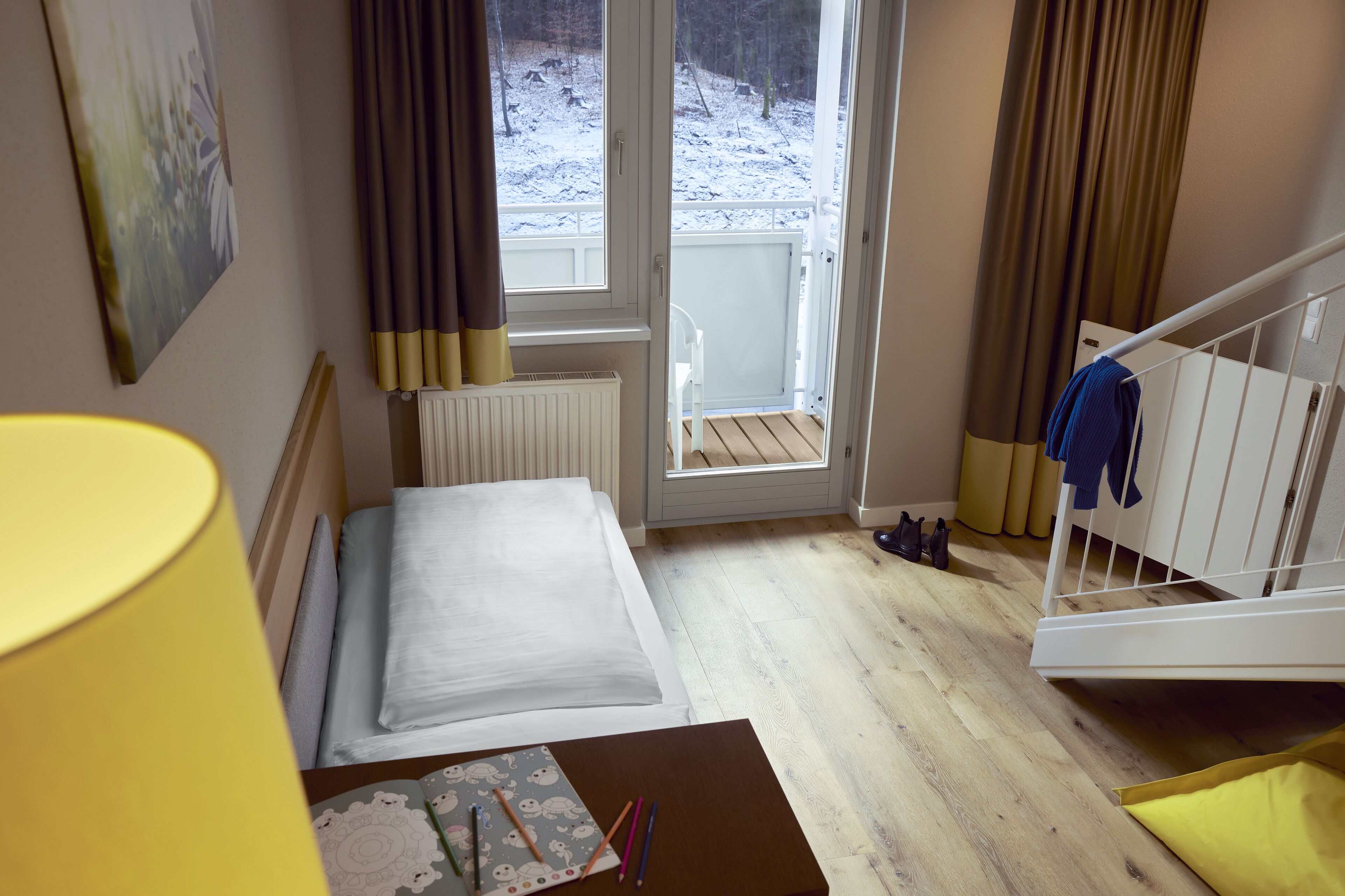 A hotel room with a bed, a chair, a small table with art supplies, a window with curtains, and a view of a snowy outdoor area.