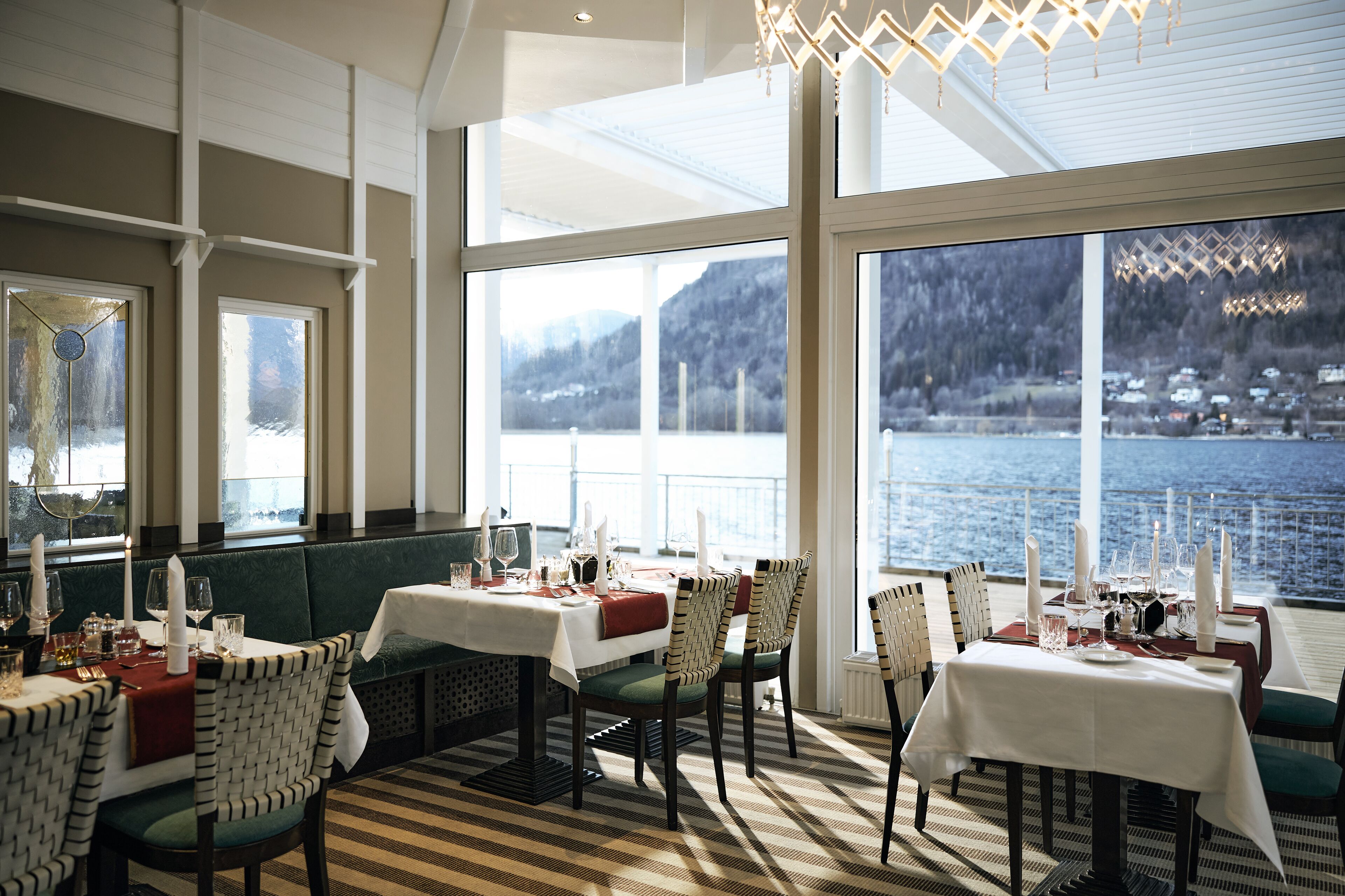 A restaurant with tables set for dining, large windows with a view of a lake and mountains, and a chandelier.
