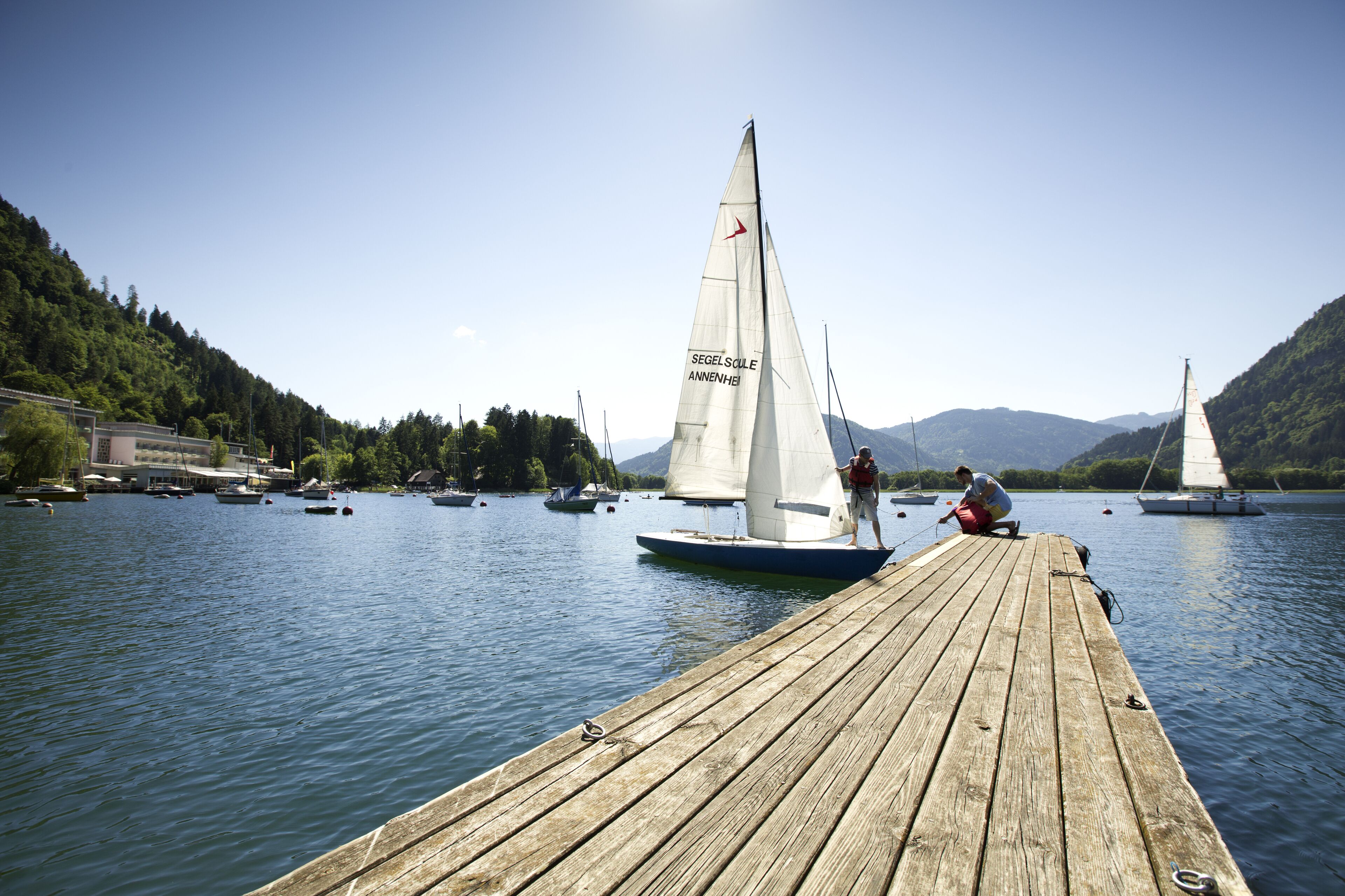 Une jetée en bois s’étendant dans un lac paisible, où l’on aperçoit des voiliers et une personne debout sur la jetée.