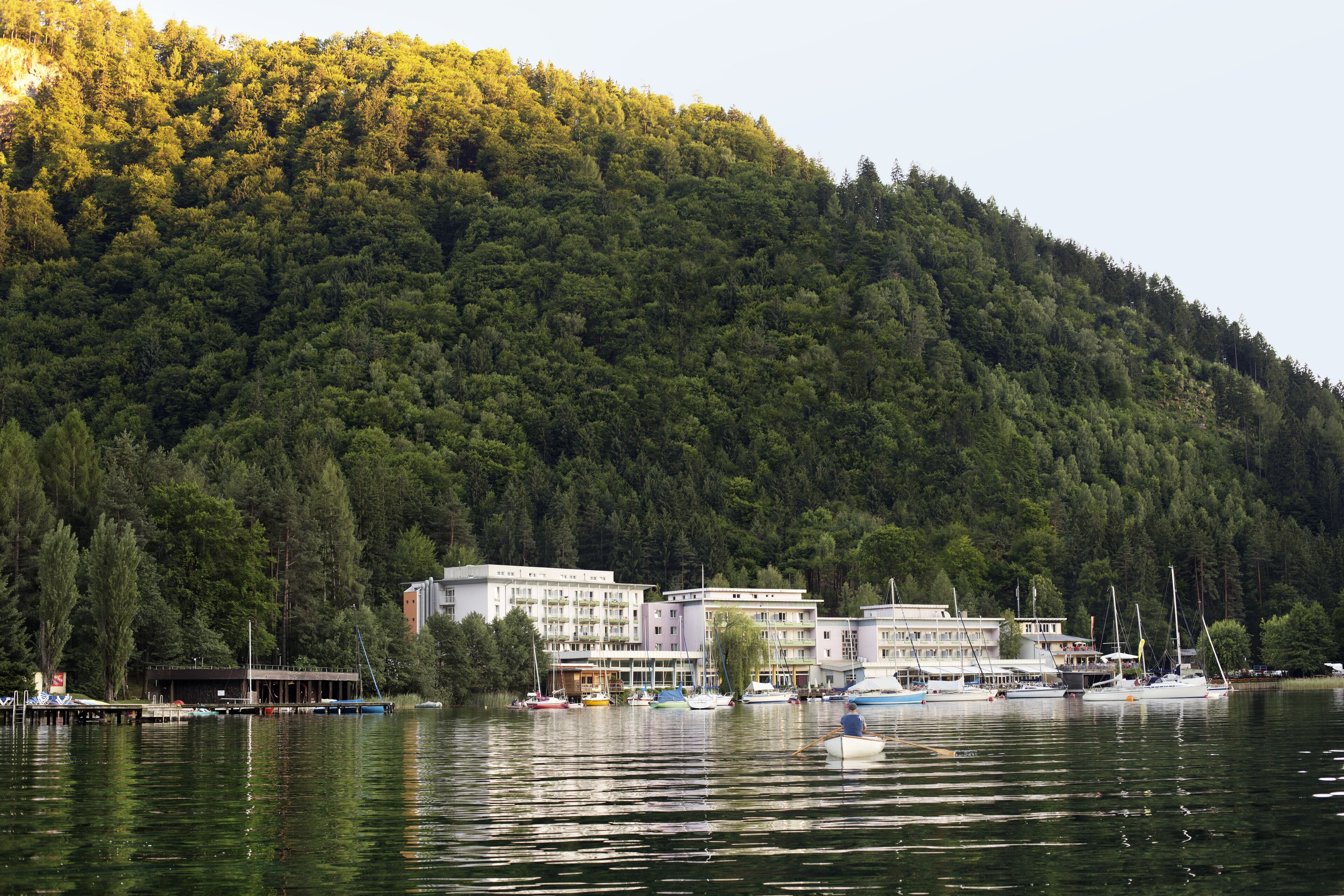 Un hotel junto al lago con varias construcciones, barcos amarrados y una ladera cubierta de bosque.