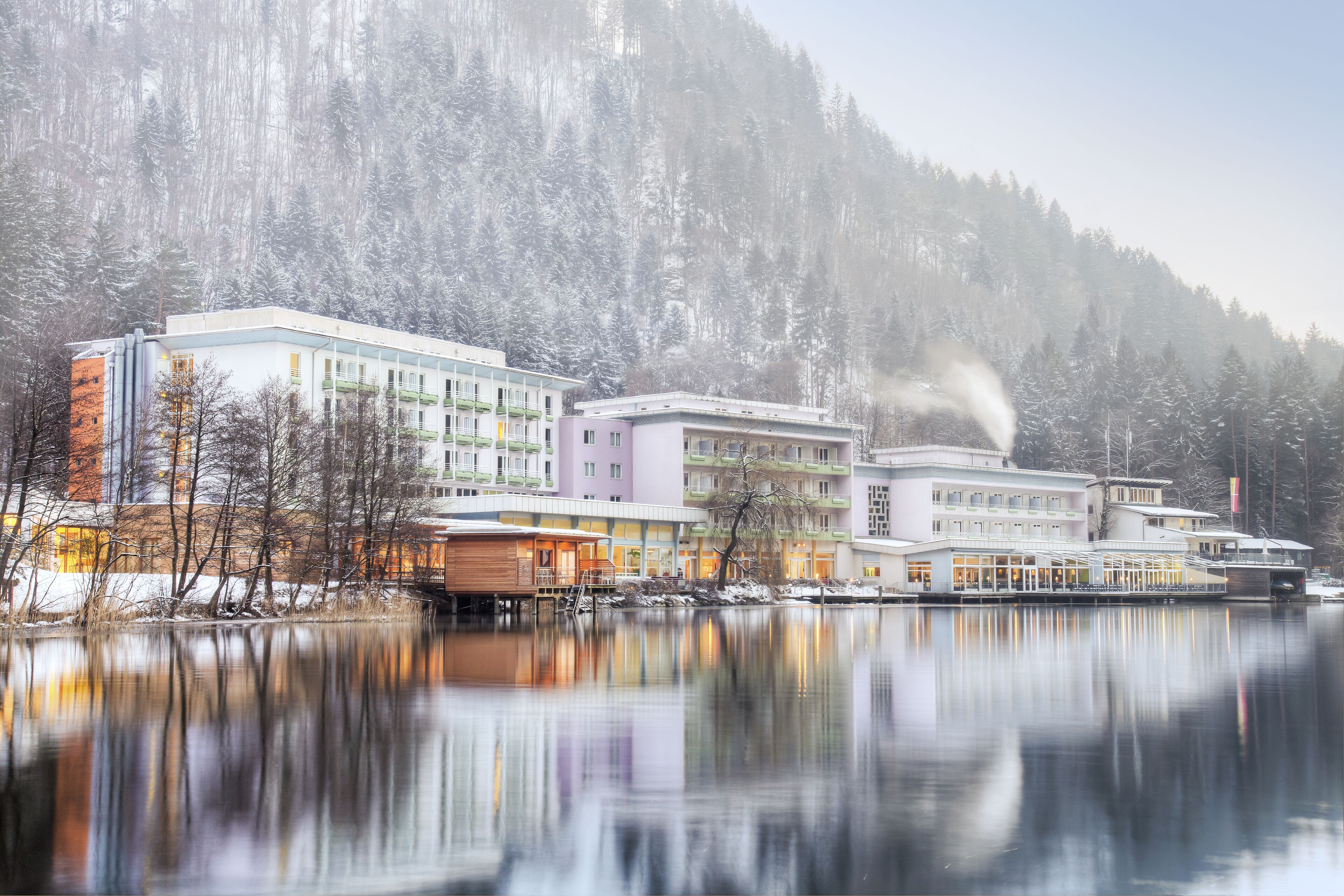 A hotel with multiple buildings by a calm lake, surrounded by snow-covered trees and a mountain.