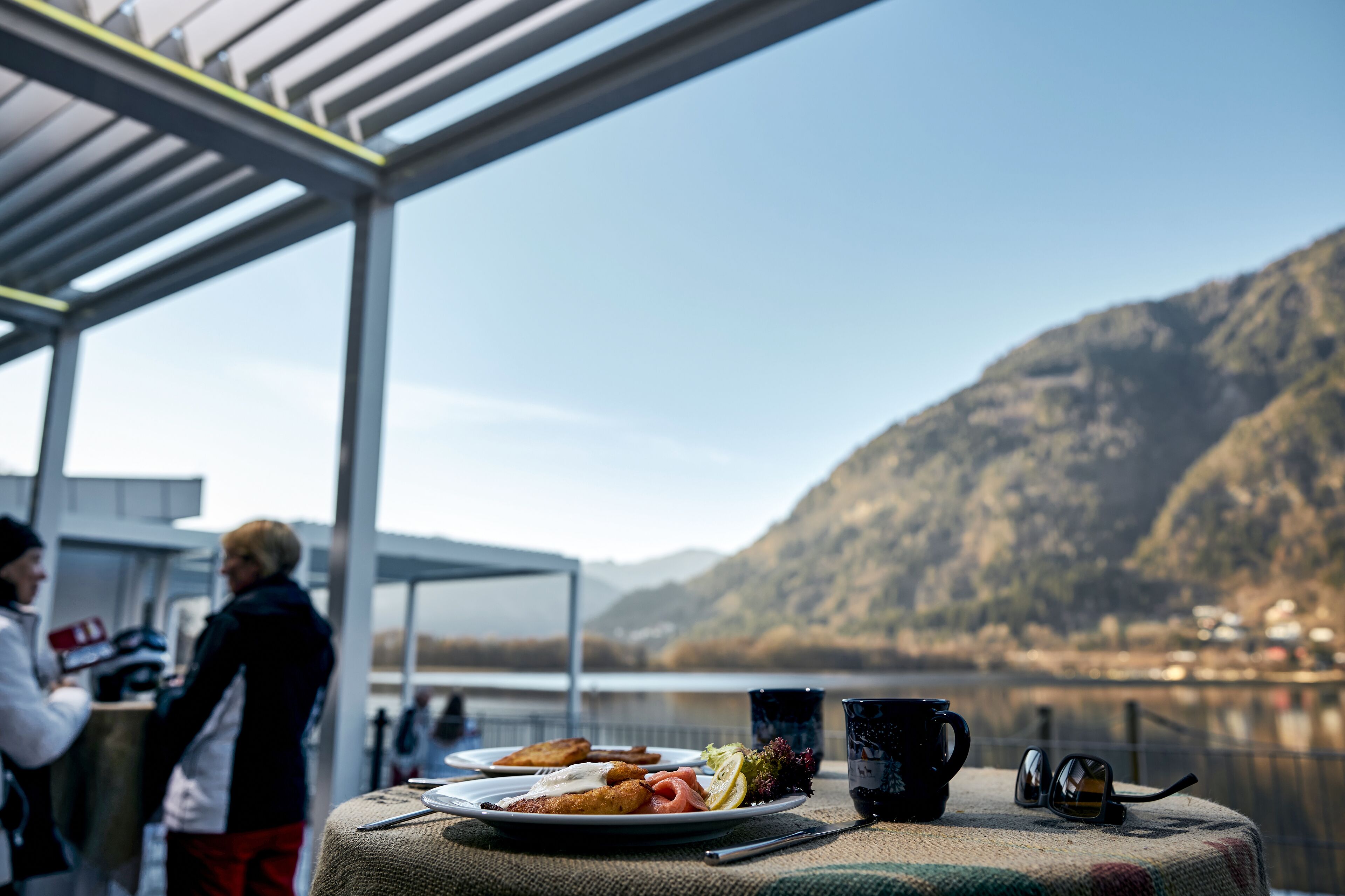 Een tafel met eten, een mok en zonnebril, met uitzicht op een meer en bergen.