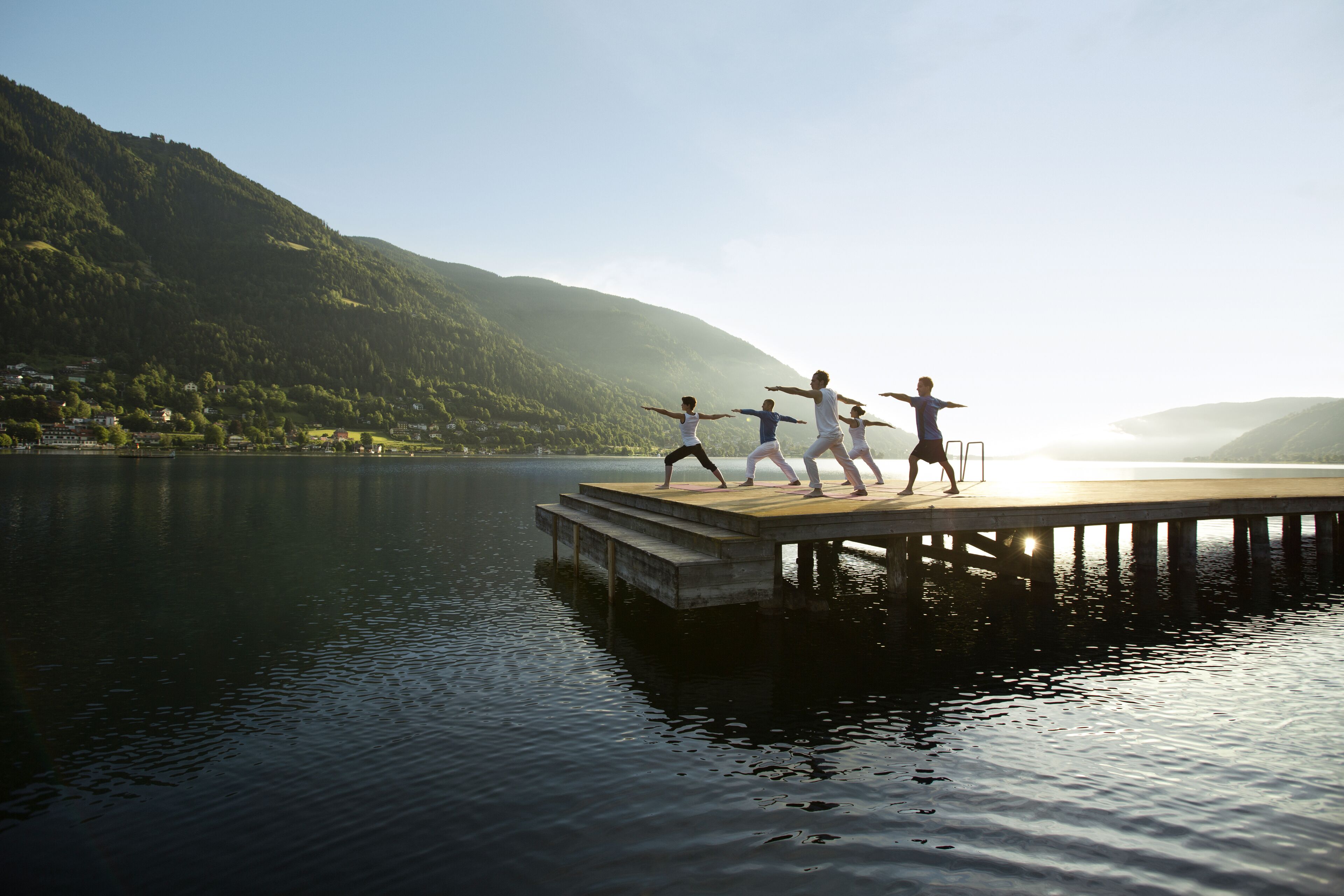 Un grupo de personas practicando yoga en un muelle de madera que se extiende sobre un lago tranquilo, rodeado de colinas verdes y exuberantes.