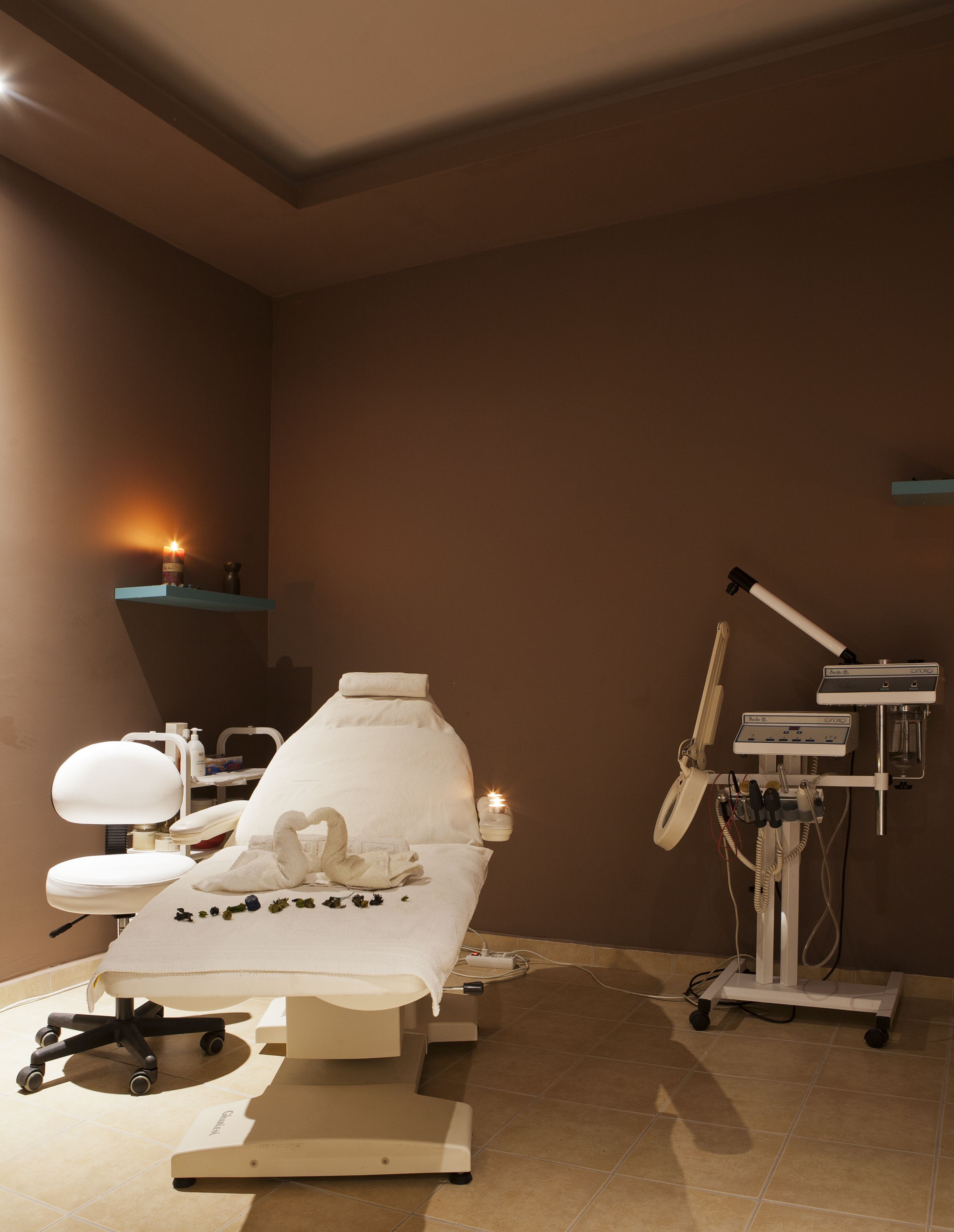 Una sala visite medica con un lettino reclinabile, un poggiapiedi, una luce d'esame montata a parete e un dispositivo medico su un carrello.