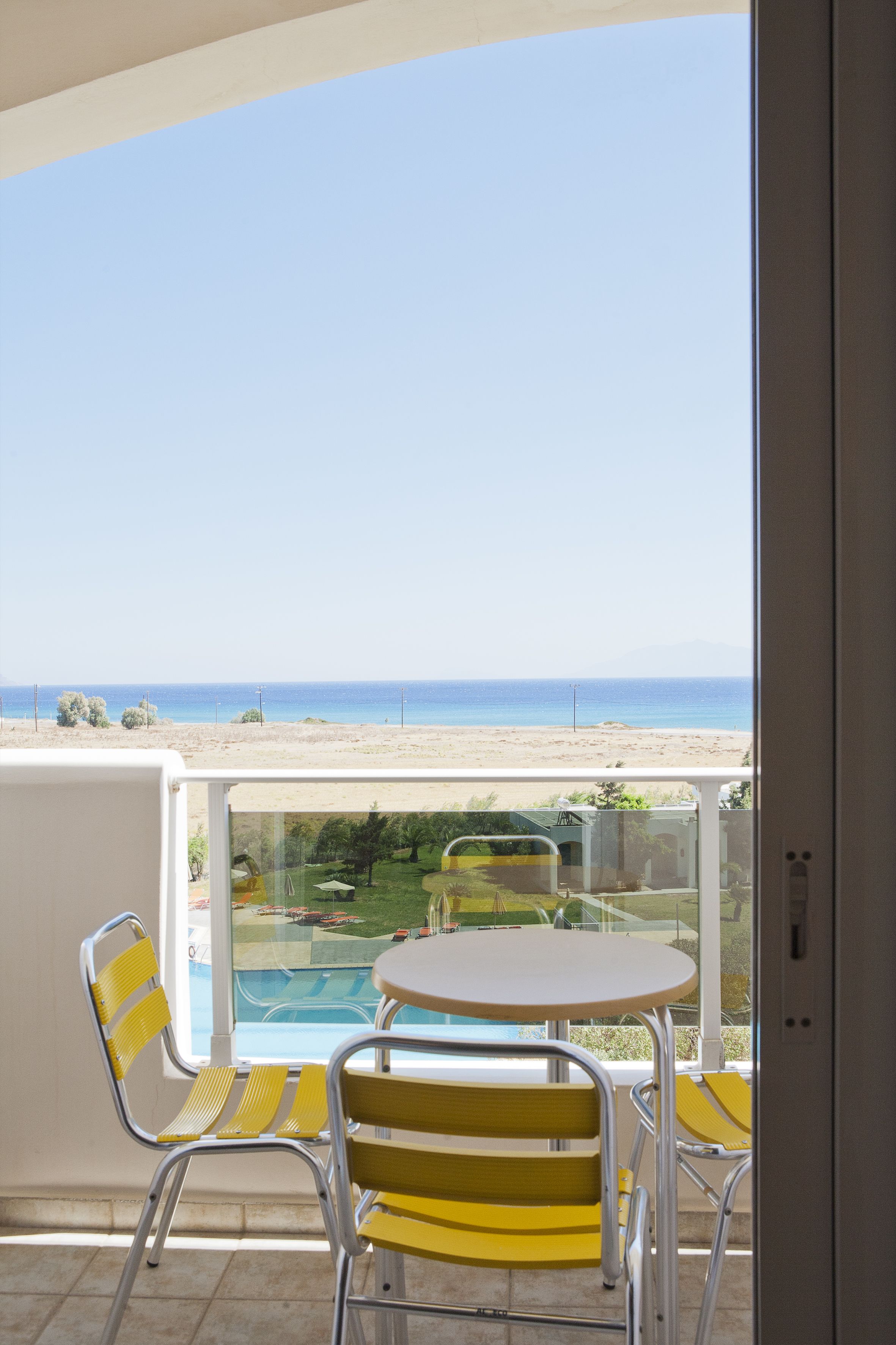Un balcón con sillas amarillas y una mesa, con vistas a una playa y una piscina.