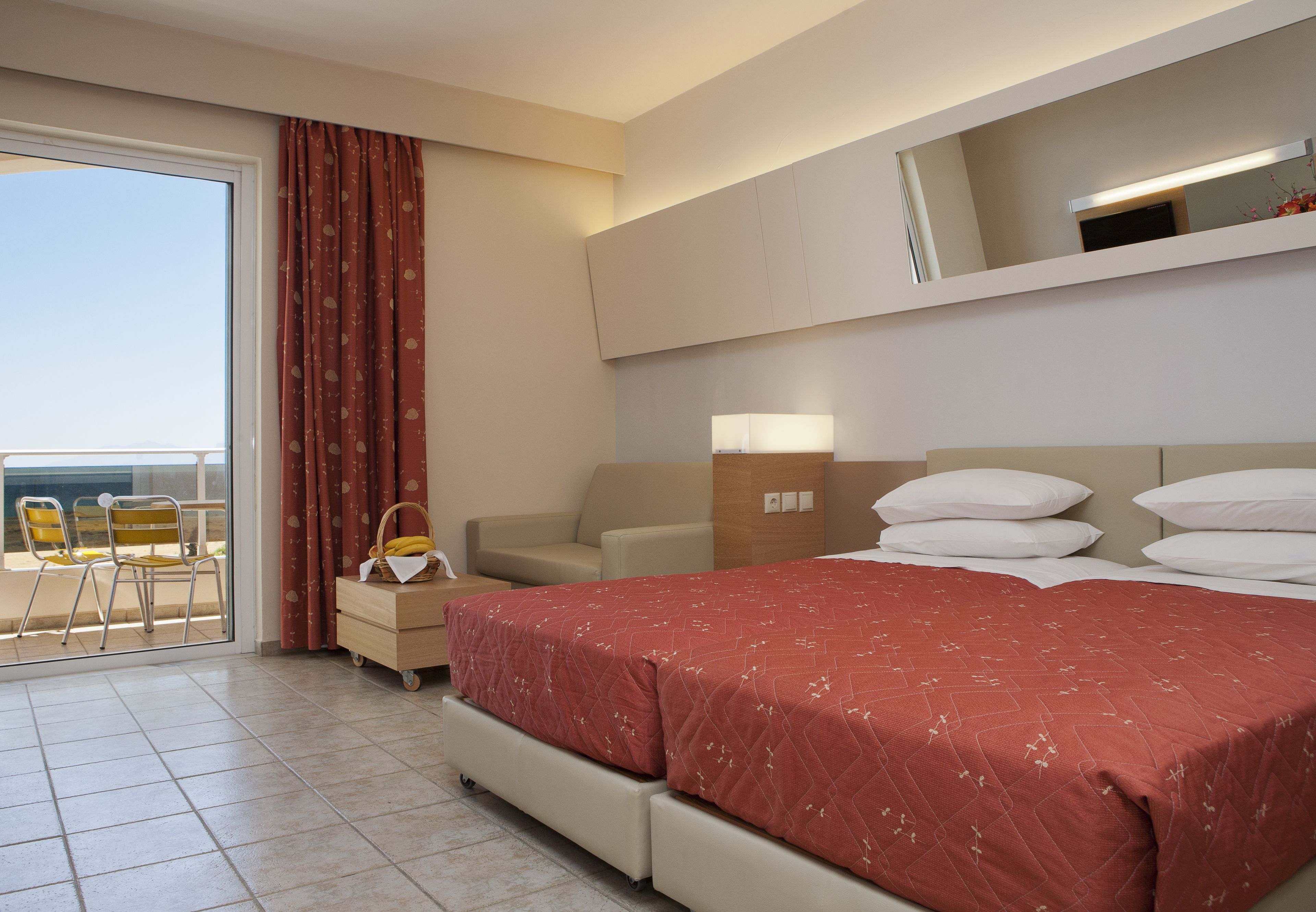 A hotel room with a bed, a sofa, a chair, a small table, a lamp, and a view of the sea through a sliding door.