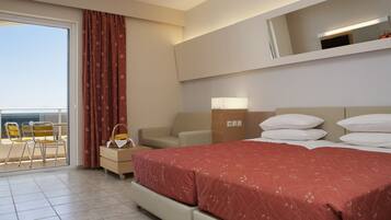 A hotel room with a bed, a sofa, a chair, a small table, a lamp, and a view of the sea through a sliding door.