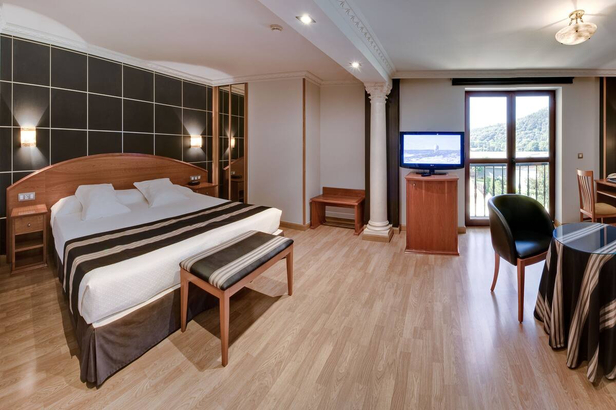A hotel room with a large bed, a desk with a chair, a TV, and a balcony with a view.
