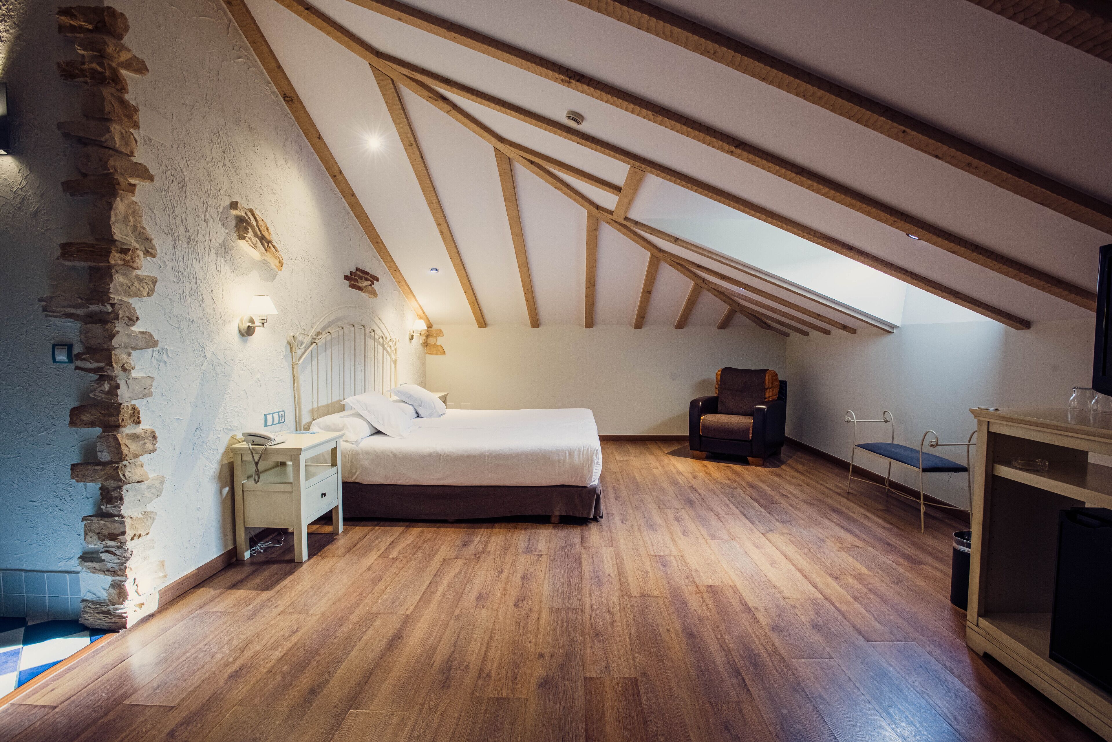 A bedroom with a sloped ceiling, wooden beams, a bed, a nightstand, a chair, and a small table.