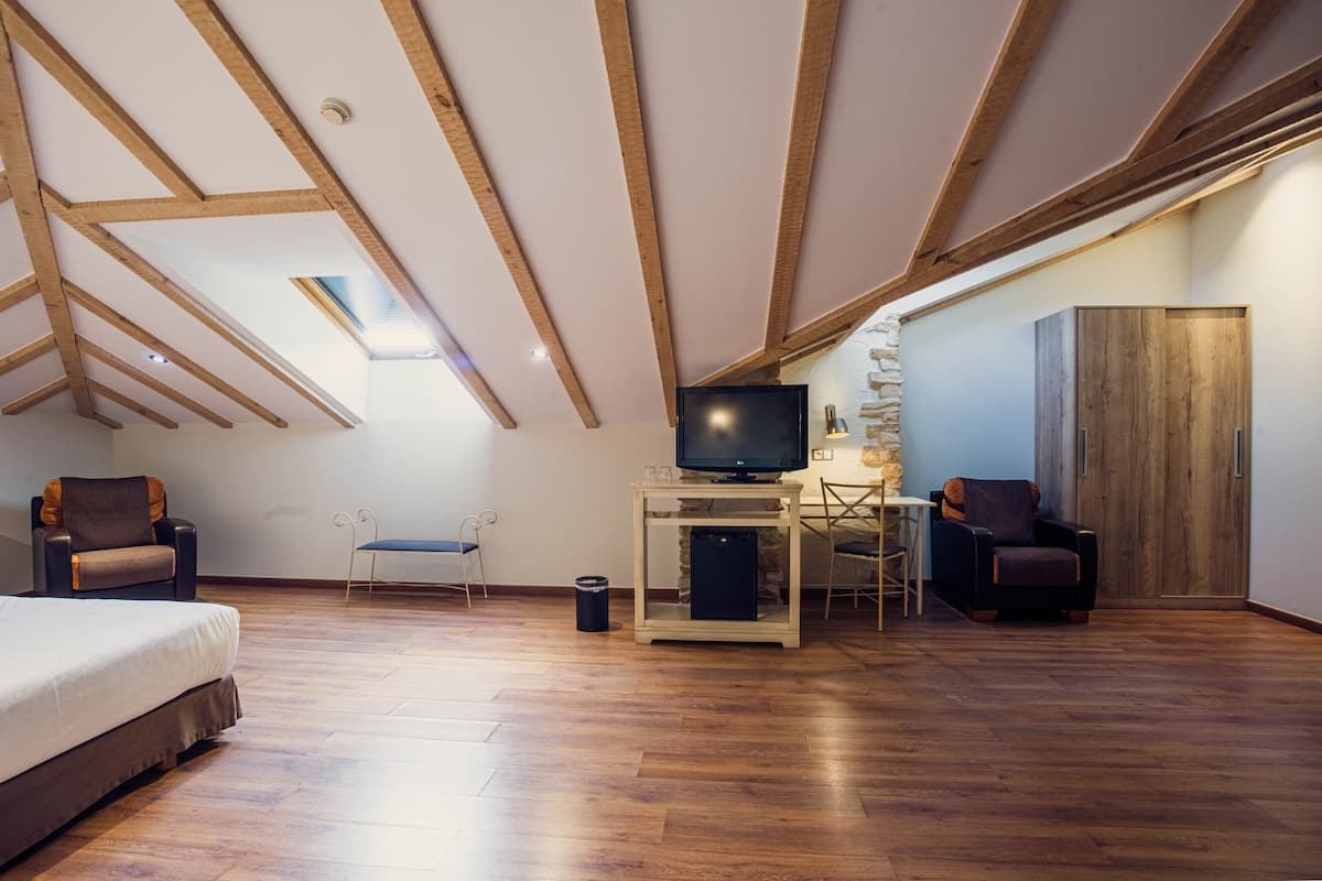 A loft-style bedroom with a sloped ceiling, wooden beams, a bed, a desk with a TV, and two chairs.