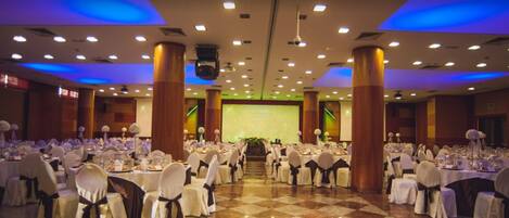 A banquet hall with round tables, white chairs, and a stage at the front.