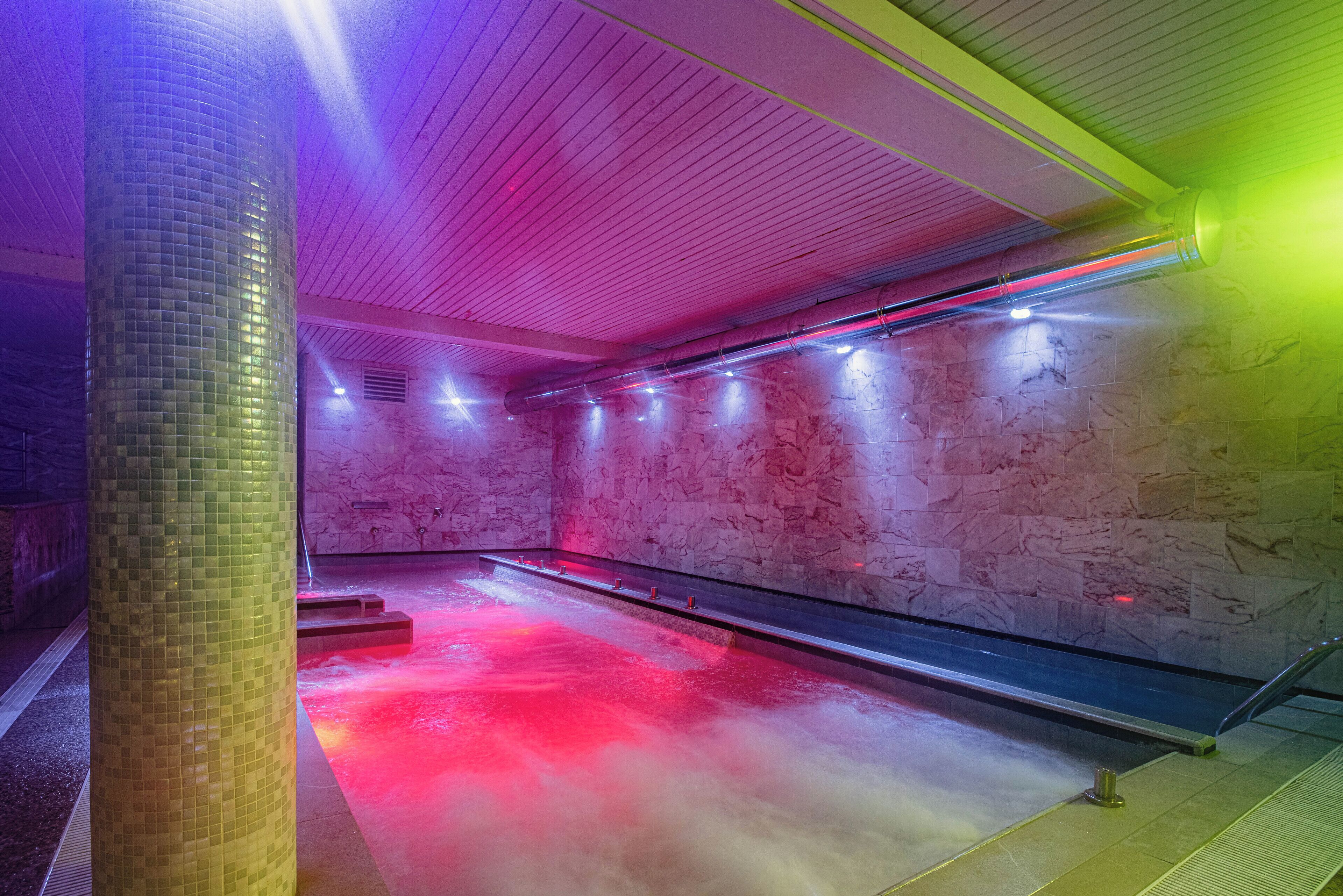 A modern indoor pool area with a tiled floor, marble walls, and a central water feature.