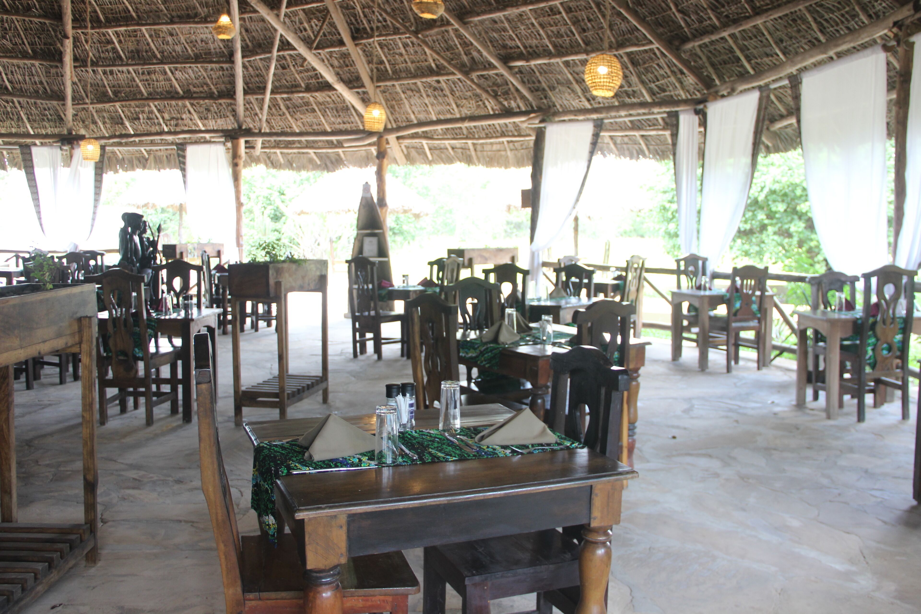 An outdoor dining area with wooden tables and chairs, a thatched roof, and hanging lanterns.