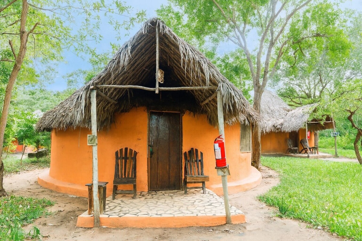A traditional hut with a thatched roof, a wooden door, and a small porch with two wooden chairs.