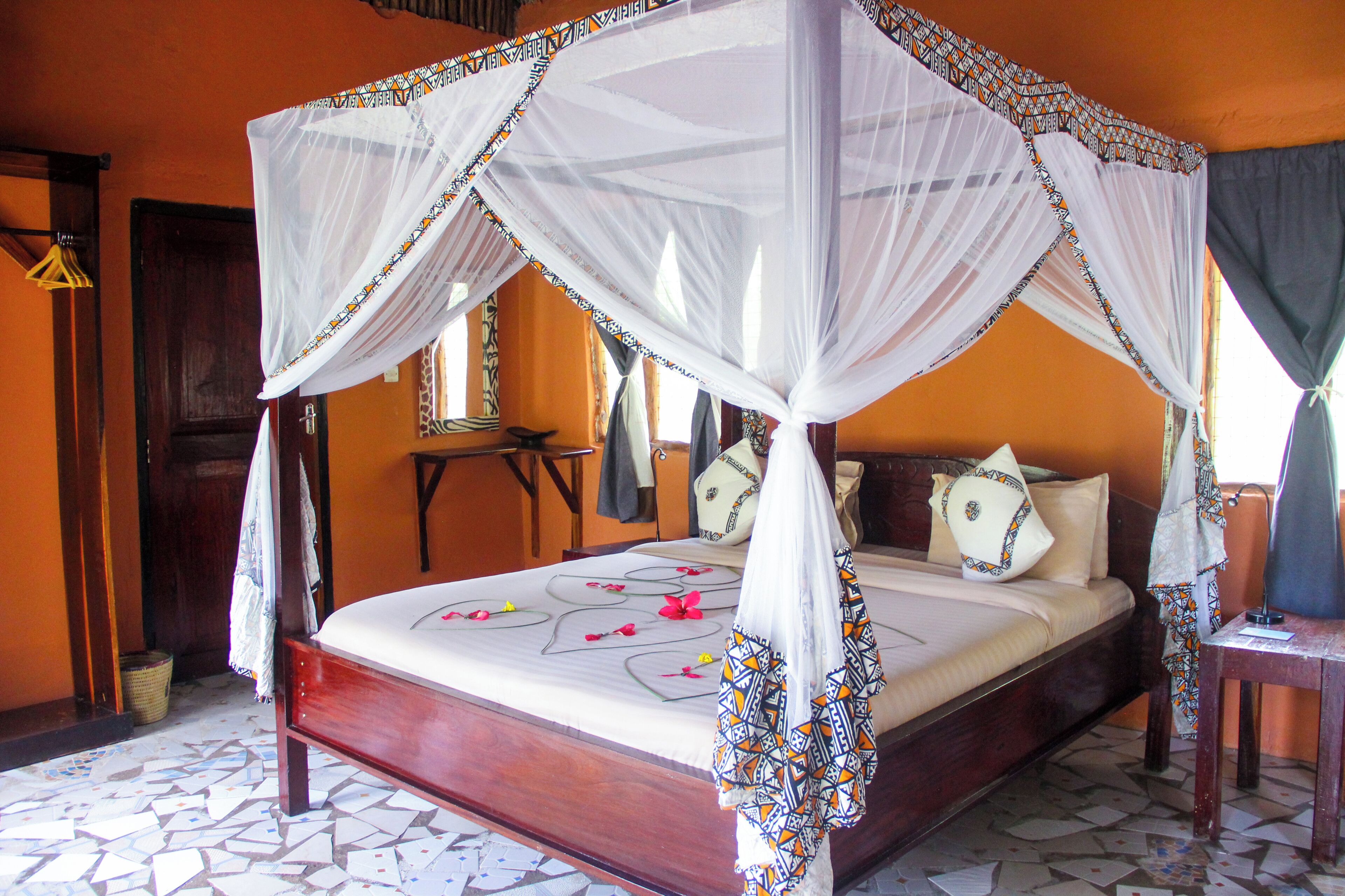 Una cama con dosel y cuatro columnas, con una colcha blanca y almohadones decorativos, un cabecero de madera y suelo de baldosas.