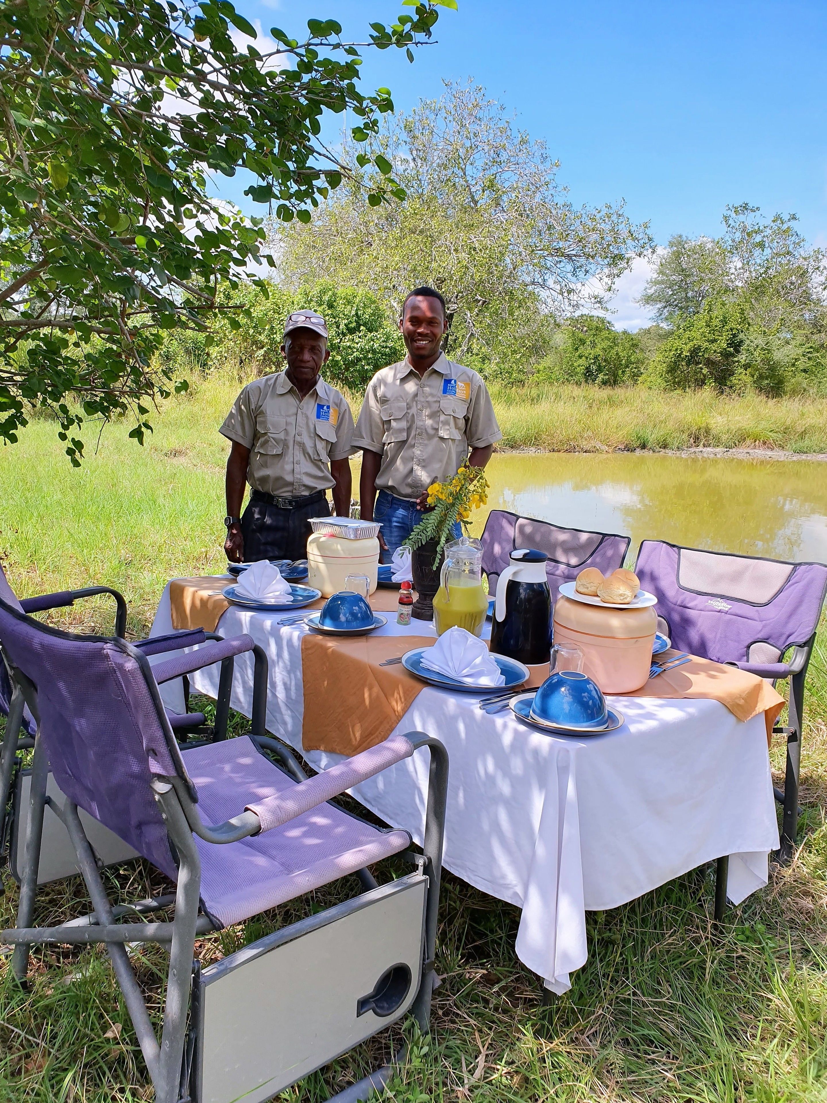 Dos hombres de pie junto a una mesa al aire libre con mantel blanco, rodeada de sillas y diversos alimentos.