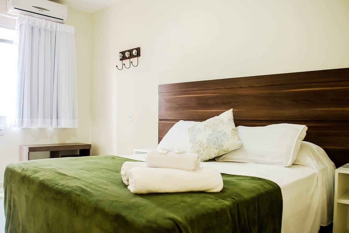 Un lit bien fait, avec une couvre-lit verte, des oreillers blancs et une tête de lit en bois. On y trouve une petite table de chevet avec une lampe, et un climatiseur fixé au mur.