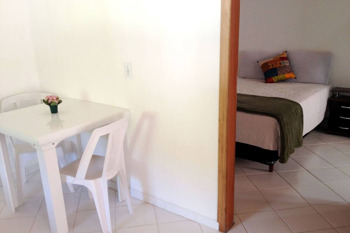 Une petite pièce propre, avec une table et une chaise blanches, un lit agrémenté d’un coussin coloré et un plancher en bois.