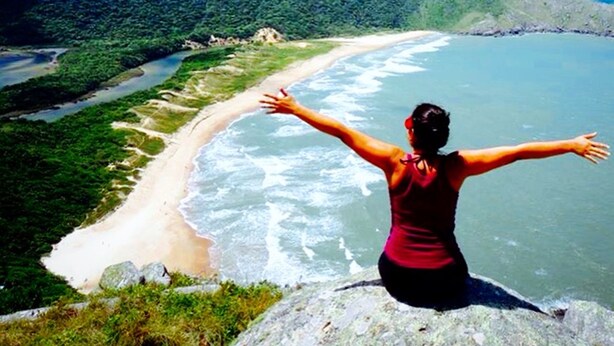 A person with arms outstretched overlooking a coastal landscape with mountains and a beach. - Pousada Pedra Rosa (Florianópolis)