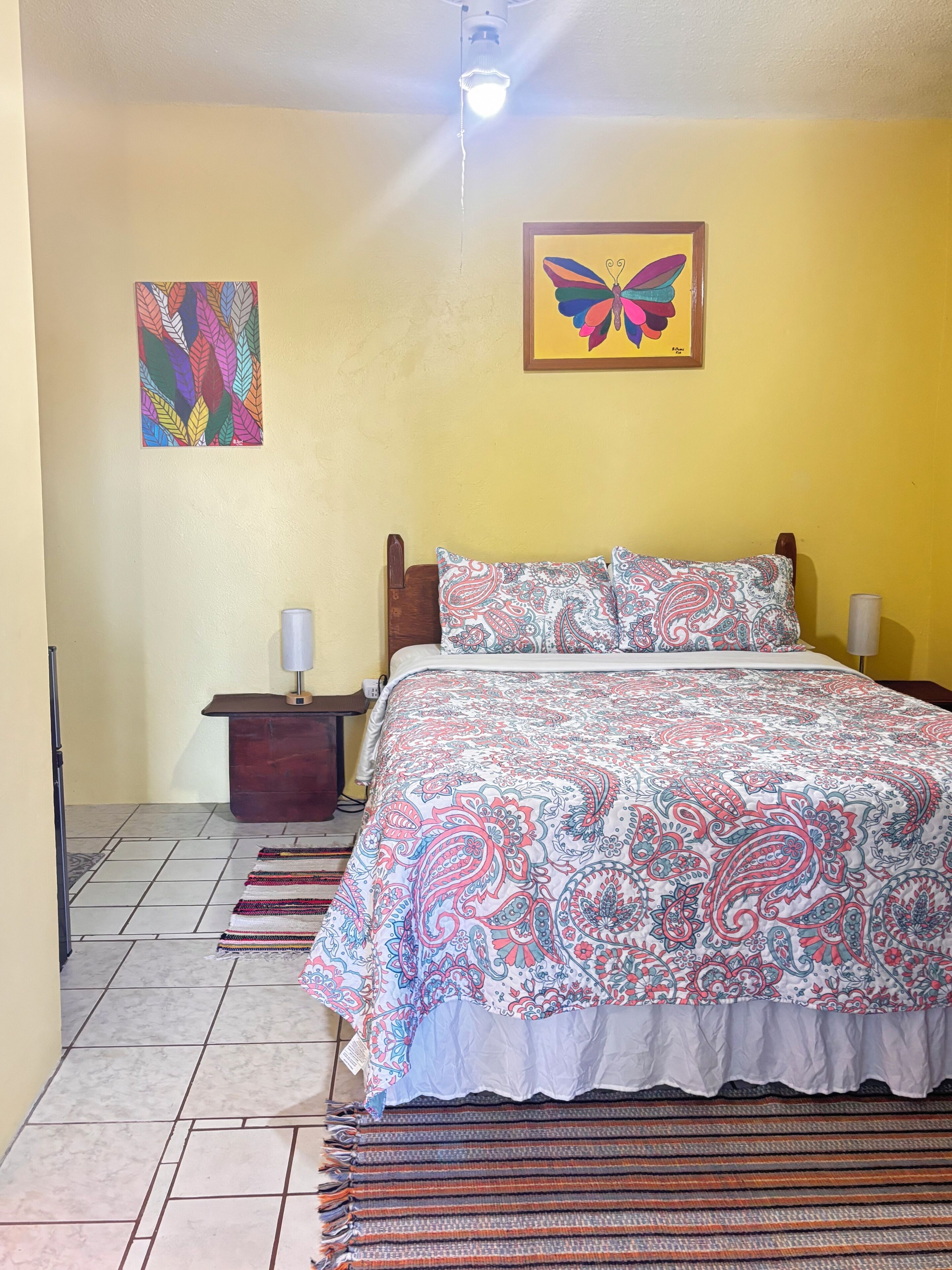 Un dormitorio con cama, mesita de noche, lámpara, ventilador de techo y dos cuadros.