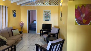 A living room with a sofa, chairs, a TV, and a ceiling fan. There is a bed in the adjacent room. - Ellen Bay Cottages (Seatons Village)