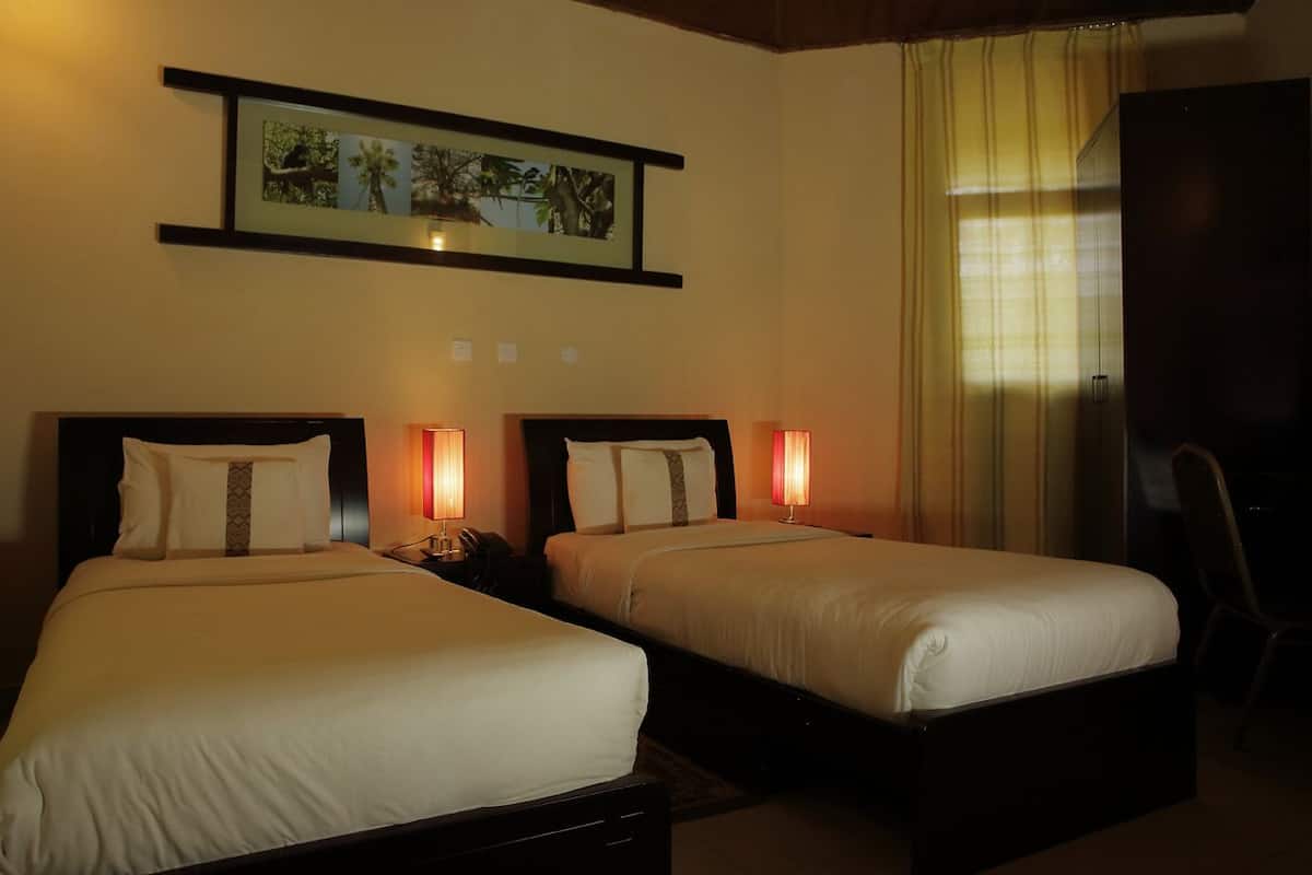 Ein Hotelzimmer mit zwei Einzelbetten, einem Nachttisch mit Lampe, einem gerahmten Bild an der Wand und einem Fenster mit Vorhängen.