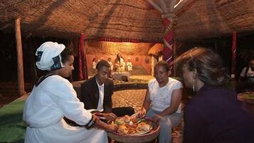 Un cadre traditionnel avec un toit de chaume, un espace central aménagé pour s’asseoir et une personne qui sert les plats.