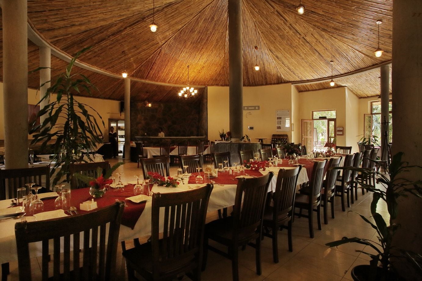 Een restaurant met ronde tafels klaar voor diners, een hoog plafond met rieten dak en potplanten.