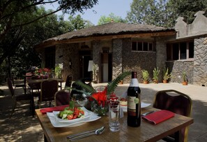Un área de comedor al aire libre con un edificio de piedra al fondo, una mesa dispuesta con ensalada, una botella de vino y un vaso de agua.