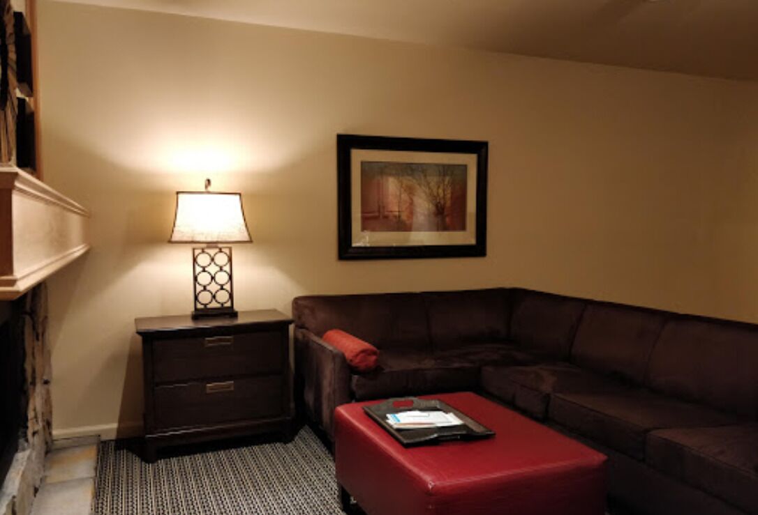 Un salon avec un canapé en cuir marron, un pouf rouge, une table d’appoint avec une lampe et un tableau encadré accroché au mur.