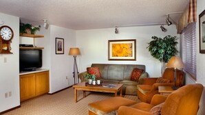 Uma sala de estar com televisão, sofá, poltronas, mesa de centro e abajur.