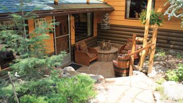 A log cabin with a stone patio, wooden furniture, and a hanging flower basket.