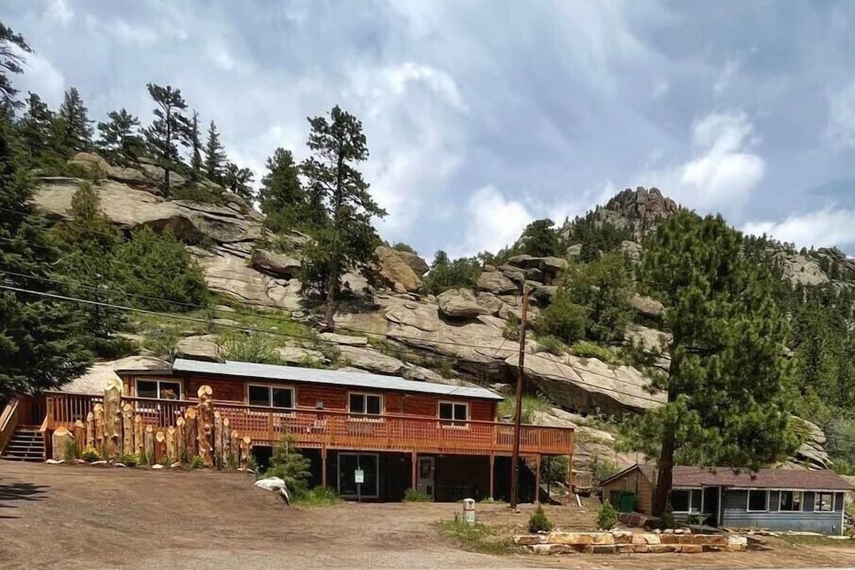 A two-story building with a wooden exterior, a porch, and a garage. It is situated on a hillside with trees and rocky terrain in the background.