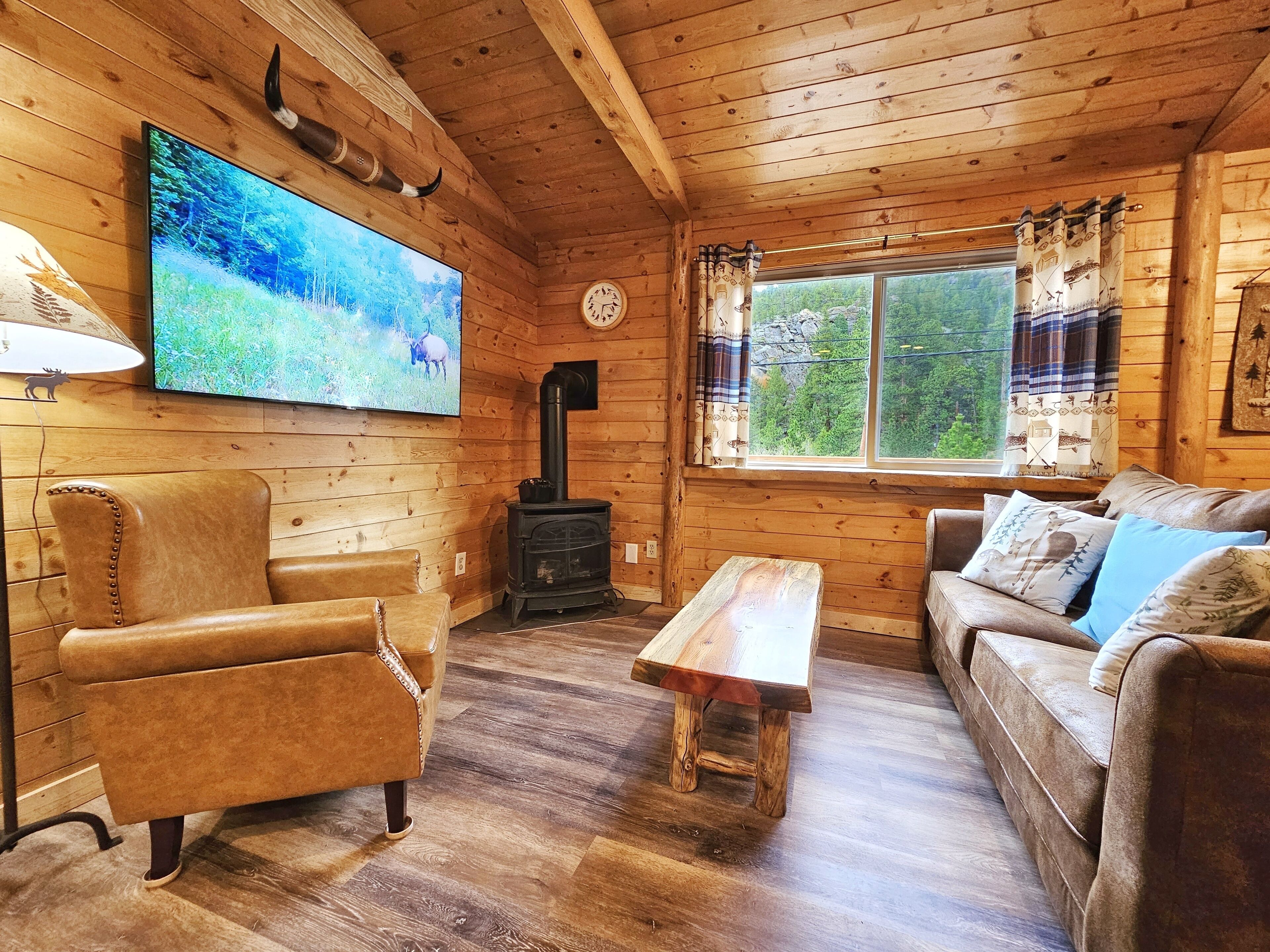 A cozy cabin interior with a wooden interior, a television mounted on the wall, a sofa, a chair, a wooden table, and a window with curtains.